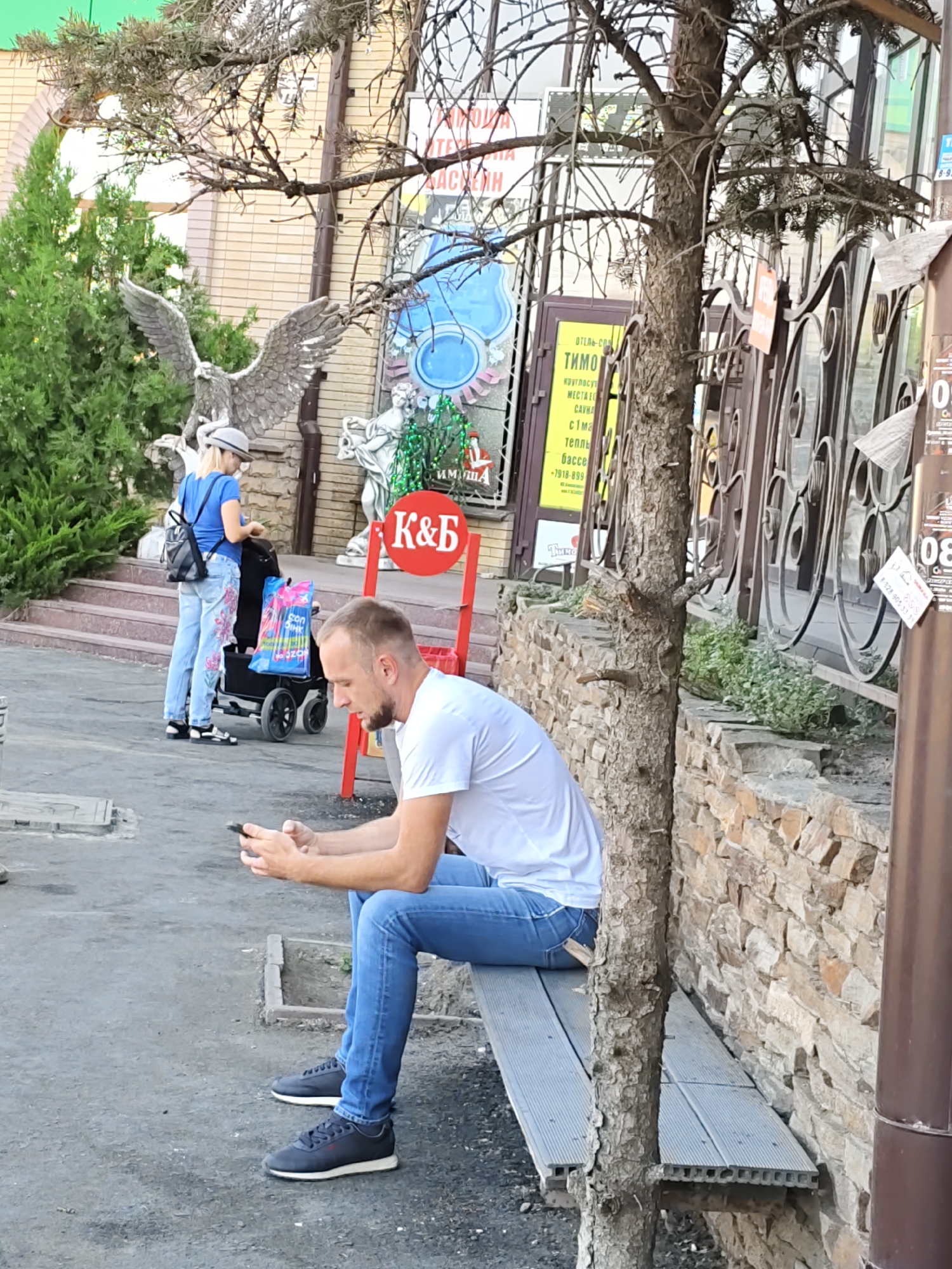 A middle-aged Caucasian man with short, blond hair and a light beard is sitting on a gray slatted bench, looking down at his smartphone, which he holds with both hands. He is wearing a white t-shirt and blue jeans. The man is positioned in the foreground, to the right of the image's center. Behind him, a light-colored stone wall with sparse greenery is visible. A thin tree trunk stands in front of the wall, partially obscuring it.
To the left of the man, a blonde woman wearing a blue t-shirt, a gray hat, and light blue jeans with a floral pattern is standing near a black stroller. She is looking down at the stroller or something within it. In the background, there are stone steps leading up to a building with beige brickwork. A red sign with white lettering that reads "K&B" is mounted on a pole near the woman. Several signs with Russian text are visible on the building and on a metal fence to the right. The environment appears to be an outdoor public space, possibly a street or park area, in Rostov, Russia.