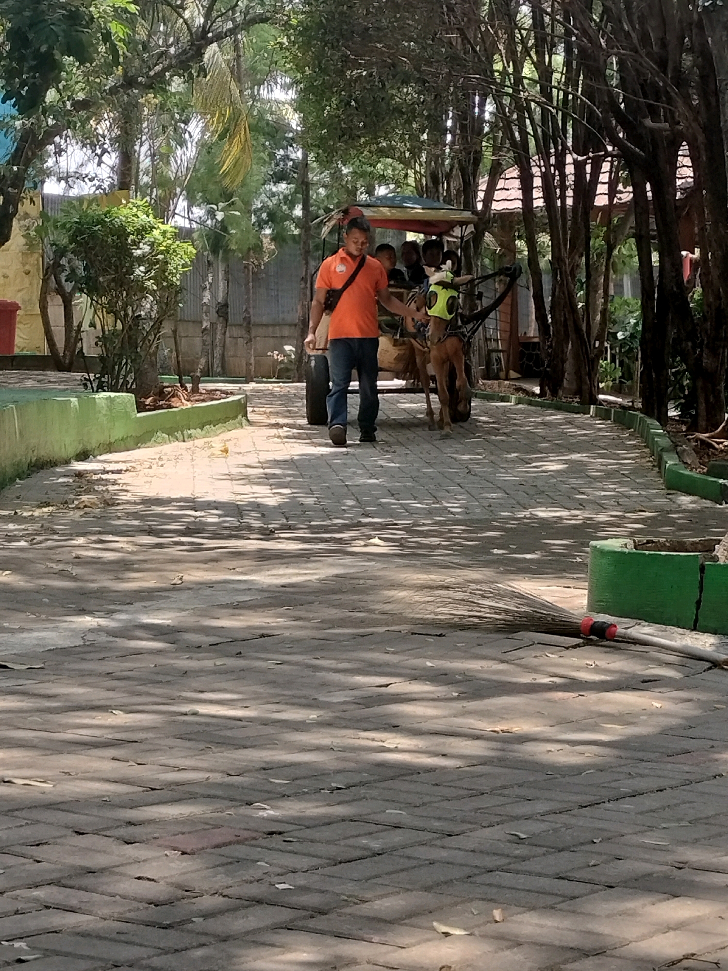 The image depicts a man in an orange shirt and blue jeans walking beside a horse-drawn carriage on a paved pathway in Depok, Indonesia. The pathway is lined with green painted borders and surrounded by trees and vegetation. The carriage appears to be carrying several passengers. A broom rests on the pathway in the foreground. The scene suggests a leisurely outdoor setting, possibly a park or tourist attraction.