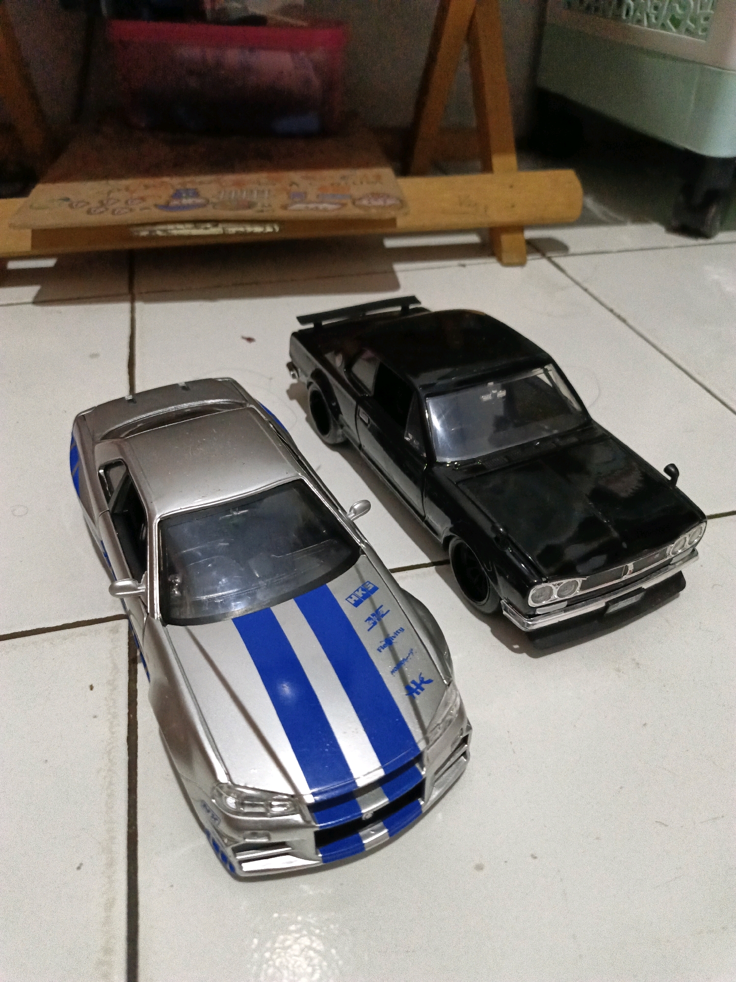 Two toy cars are positioned on a tiled floor. The car in the foreground is silver with blue racing stripes and appears to be a Nissan Skyline GT-R. The car behind it is black and resembles a classic Japanese sedan, possibly a Nissan Skyline GT-X. In the background, a wooden easel supports a plank with various stickers or decals. To the right, a portion of a light green plastic storage bin with wheels is visible. The overall setting suggests an indoor environment, possibly a domestic space. The image was taken in Keleyan, Indonesia.