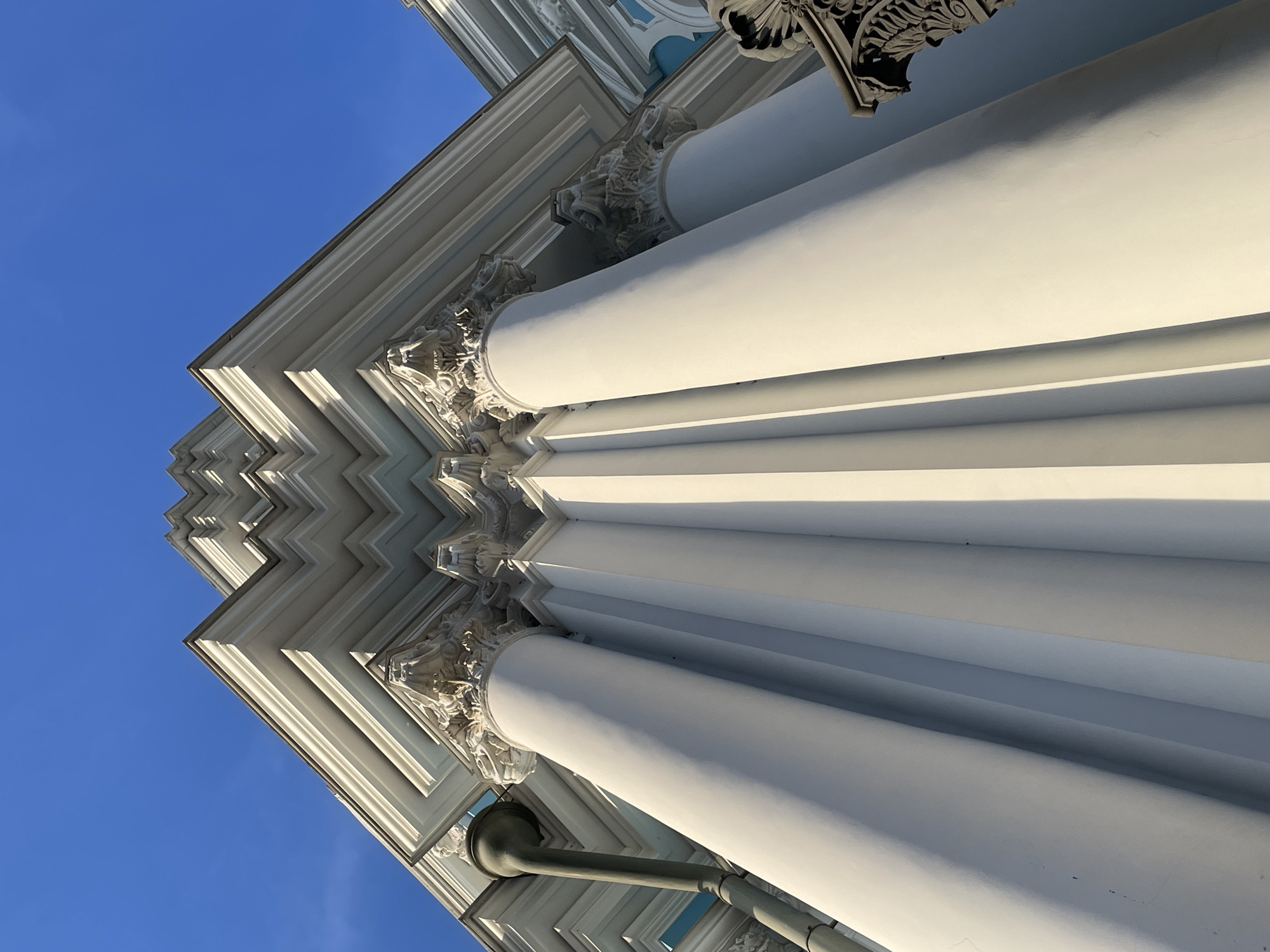 A low-angle, worm's-eye view captures the architectural details of a building's corner against a clear, bright blue sky in Unknown, Unknown. The scene features multiple large, smooth, white or off-white columns with ornate, possibly Corinthian-style, capitals. Above the capitals, a complex, stepped entablature or cornice system recedes upwards, creating a tiered, zig-zag pattern. Strong sunlight illuminates the facade, casting distinct shadows that emphasize the three-dimensional relief of the columns and the multi-layered upper structure. A dark grey or olive-green downspout is visible vertically on the far left of the building. On the far right, a dark, carved architectural detail is partially visible. Sections of the building, particularly in the background, show subtle hints of light blue or turquoise paint, contrasting with the dominant white and the vibrant blue sky. No people or actions are present.