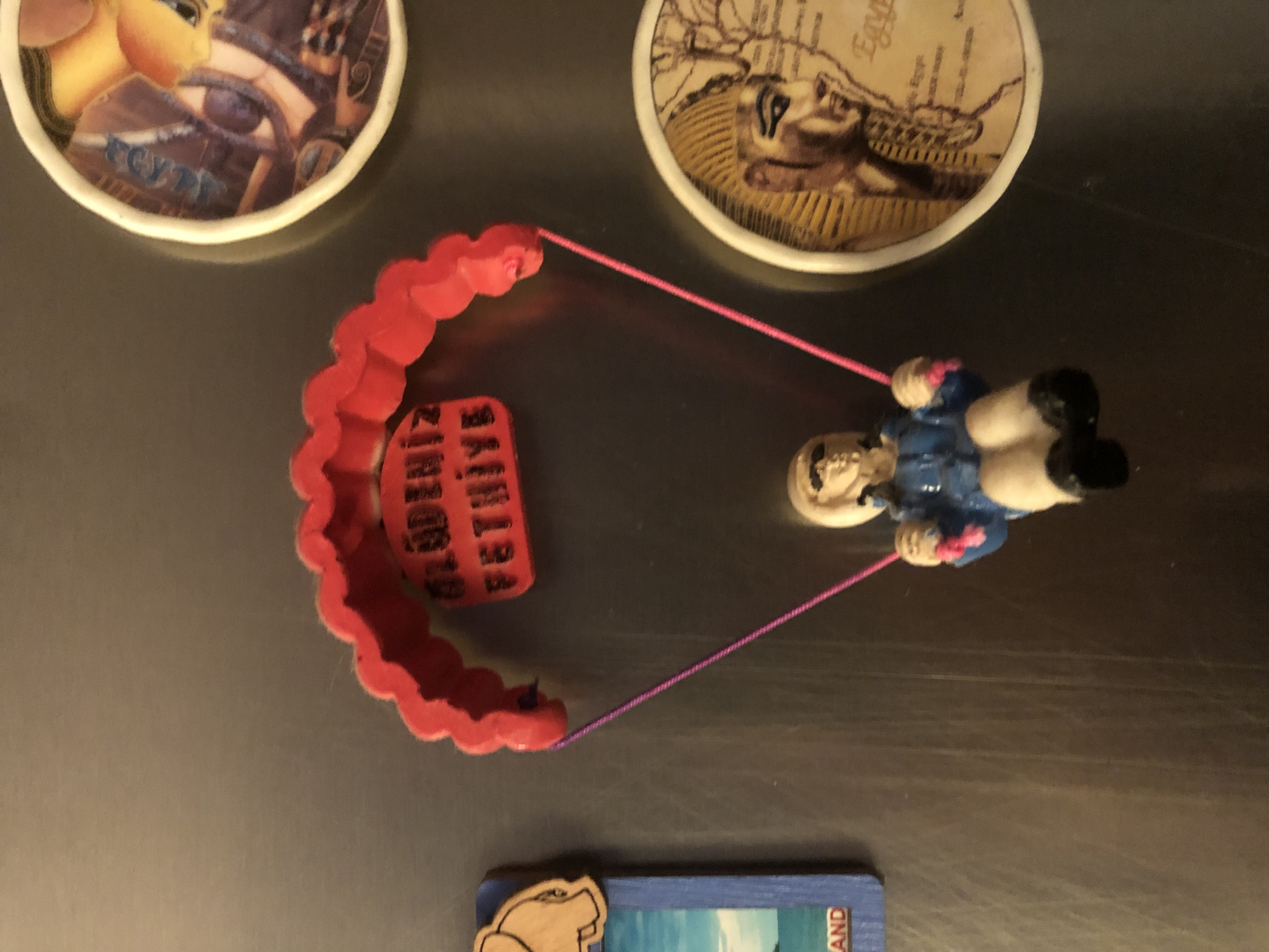 A paragliding figurine hangs from a red parachute with "ÖLÜDENIZ FETHIYE" written on a red tag. The figurine is wearing a blue jacket, tan pants, and black shoes. It is suspended by pink cords. To the left of the figurine is a rectangular magnet with a depiction of an elephant and the word "AND." To the upper right is a circular magnet with an image of a pharaoh and the word "EGYPT." Below that is another circular magnet featuring a detailed drawing of a pharaoh's head and hieroglyphics. The background appears to be a metallic surface, possibly a refrigerator. There is no indication of the city of Arfa, Russia.