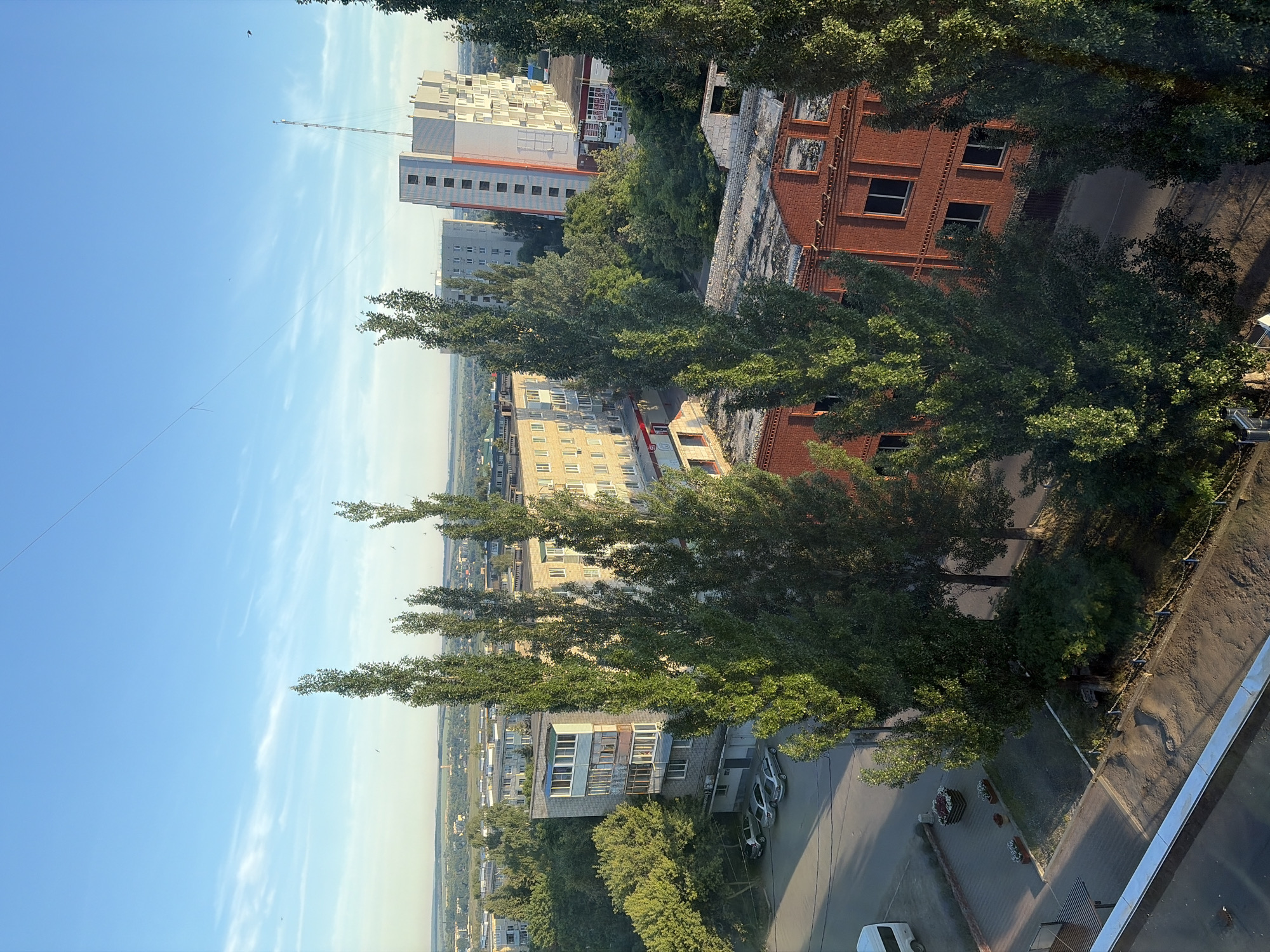 The image depicts a view of an urban environment in Skopin, Russia, characterized by a mix of residential and commercial buildings, as well as significant tree cover. The scene is captured under a clear blue sky with a few wispy clouds.
In the foreground, tall, slender trees with dense green foliage dominate the view. To the left, a white van is parked on a paved area next to a building with a brick lower section and a lighter upper facade. Several cars are parked in a row further down the street.
Mid-ground structures include multi-story apartment buildings, some with balconies and visible windows. One prominent building is white with orange accents and a tall antenna on its roof. Adjacent to it is a shorter, light-colored building with multiple windows. There's also a brick building with numerous dark-framed windows visible in the middle right of the frame. A flatter, red brick building with open window frames is also visible in the lower right portion of the image.
The background reveals a more distant urban landscape with more buildings and trees stretching towards the horizon, suggesting a moderately sized city. The overall impression is a daytime scene, likely in the late afternoon or early morning due to the angle of the sunlight casting shadows and illuminating the scene. No people are visible in the image. The environment appears to be a well-established urban area with a notable presence of mature trees interspersed among the architecture.