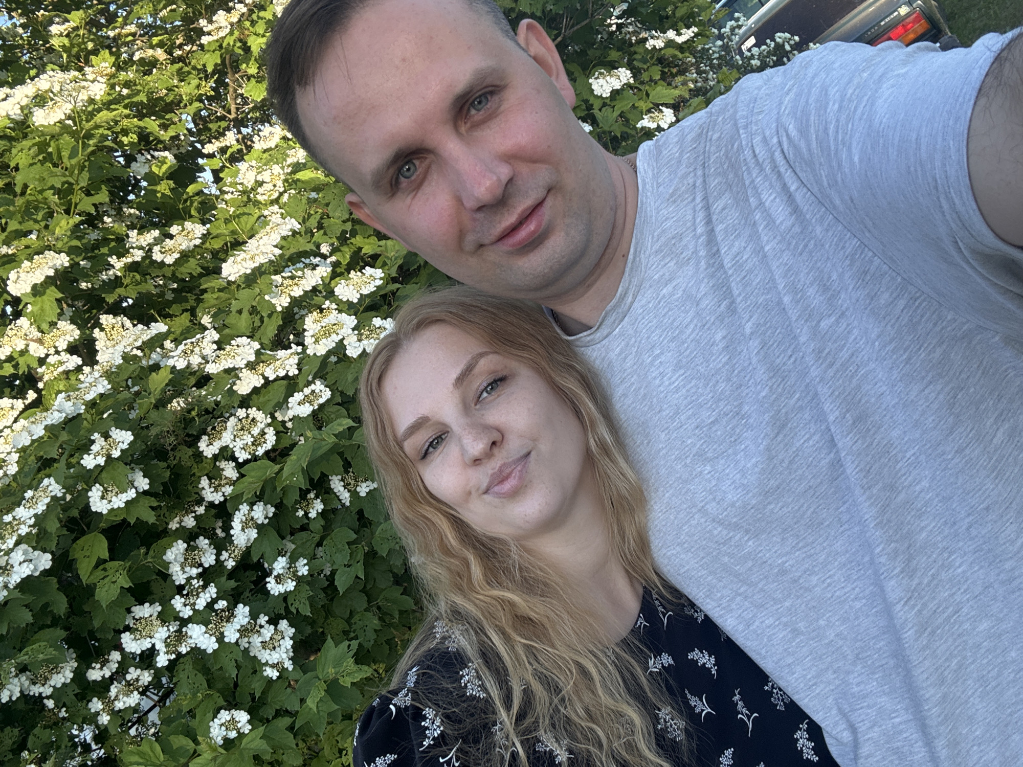An overhead, slightly tilted selfie captures a man and a woman posing outdoors in Lepilovo, Russia. The man, positioned on the right (viewer's left), has short dark hair, blue-grey eyes, and wears a light grey marled t-shirt with a thin chain visible at his neck. He exhibits a slight smile. The woman, leaning against his left side with her head resting on his shoulder, has long wavy blonde hair, fair skin with visible freckles across her nose and cheeks, and green eyes, also displaying a slight smile. She is wearing a dark-colored top with a white floral or botanical pattern. Behind the couple, a dense bush or small tree is covered in numerous clusters of small white flowers with yellowish centers and green leaves, some showing minor insect damage. In the far right background, the rear section of a dark-colored car, identifiable as a Lada by its taillights and a partially visible "LADA" emblem, is present, suggesting a roadside or outdoor parking area under natural daylight.