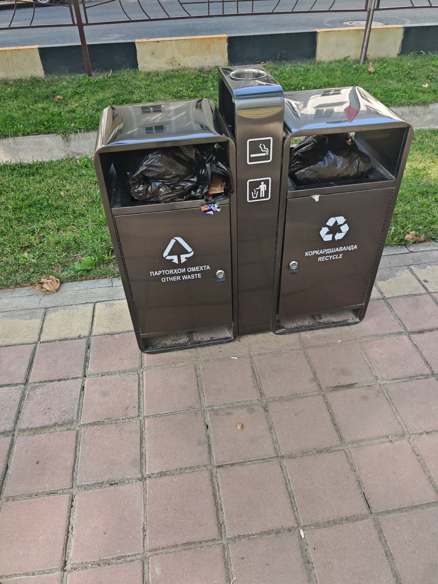 An outdoor medium shot shows a dark brown, dual-compartment trash receptacle situated on a paved sidewalk adjacent to a grassy area. The receptacle has two openings at the top, one for general waste and another with a metal insert, likely for cigarette butts. Each compartment has a door at the front with a small silver lock. The left compartment displays a white symbol of a recycling triangle above the text "ПАРТОБХОИ ОМЕХТА OTHER WASTE". The right compartment has two white icons on its front: one depicting a smoking pipe and another of a person with a broom and dustpan. Below these icons, the text "КОРКАРДШАВАНДА RECYCLE" is visible, accompanied by a recycling symbol. Both compartments appear to contain black trash bags. In the background, a fence separates the sidewalk from a paved road and a building's reflection can be seen on the curved surface of the right compartment. The ground is covered with a pattern of square concrete pavers, and a few dry leaves are scattered on the grass.