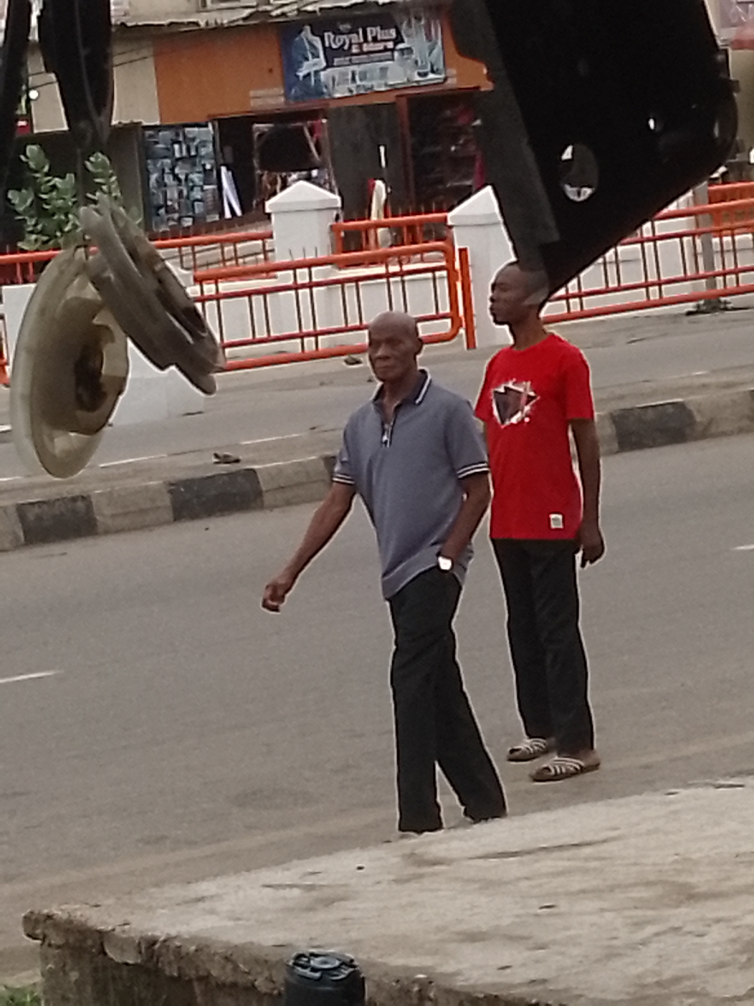 The image shows two African men walking on a city street in Lokoja, Nigeria. The man in the front wears a grey polo shirt and black pants with a wristwatch on his left wrist. The man behind him is wearing a red t-shirt with black pants and sandals. An orange barricade borders the street, and a white building with a sign that says "Royal Plus Store" can be seen in the background. There is also a heavy, dark, indistinct object looming in the foreground above the men.