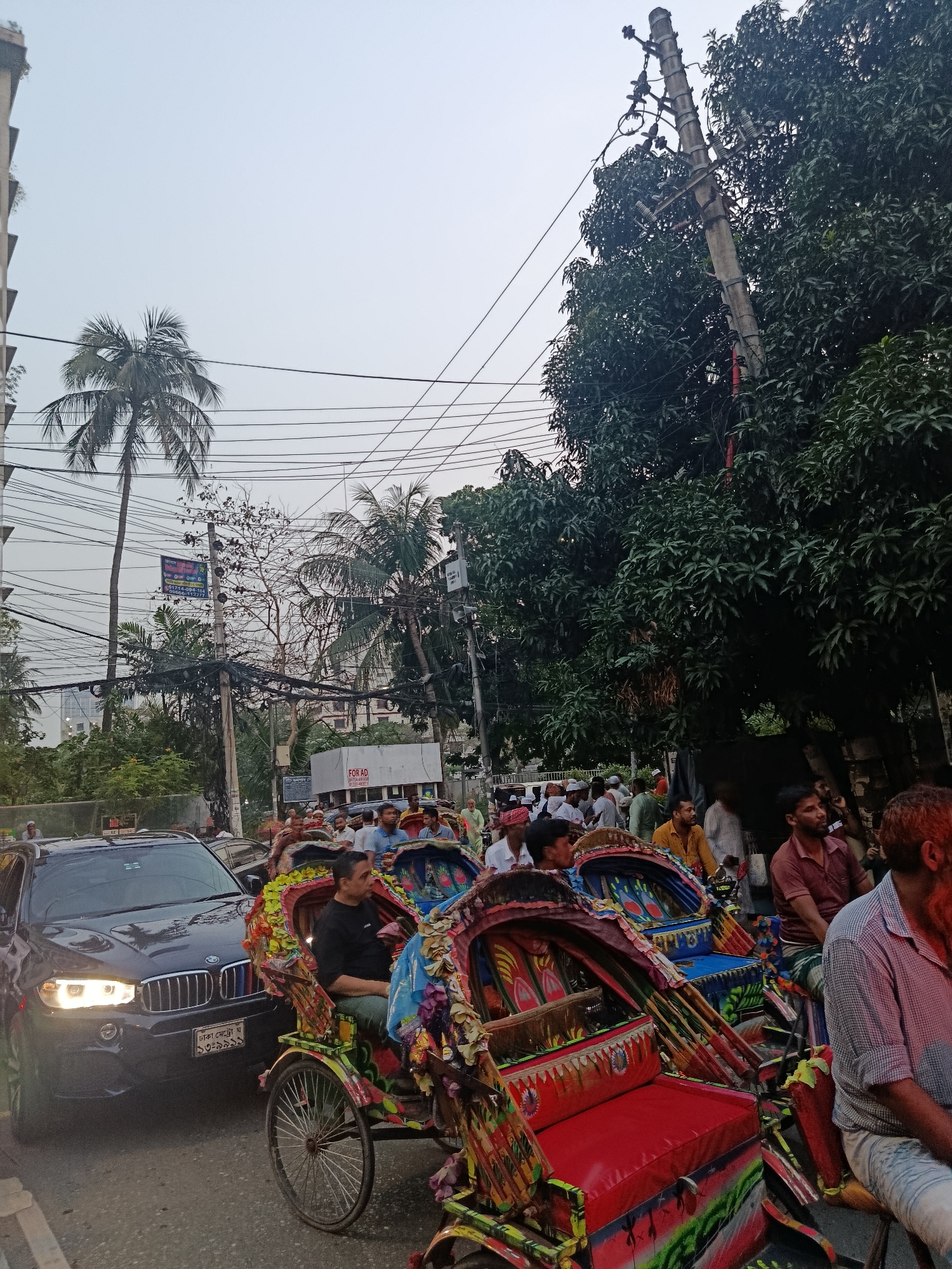 The image shows a crowded street scene in Dhaka, Bangladesh. Several brightly decorated cycle rickshaws, each carrying a passenger, are in the foreground. A black BMW SUV is visible to the left, its headlights on. A dense crowd of people, primarily men, are walking or standing along the street. Buildings and trees line the street, with power lines running overhead. The sky is overcast.