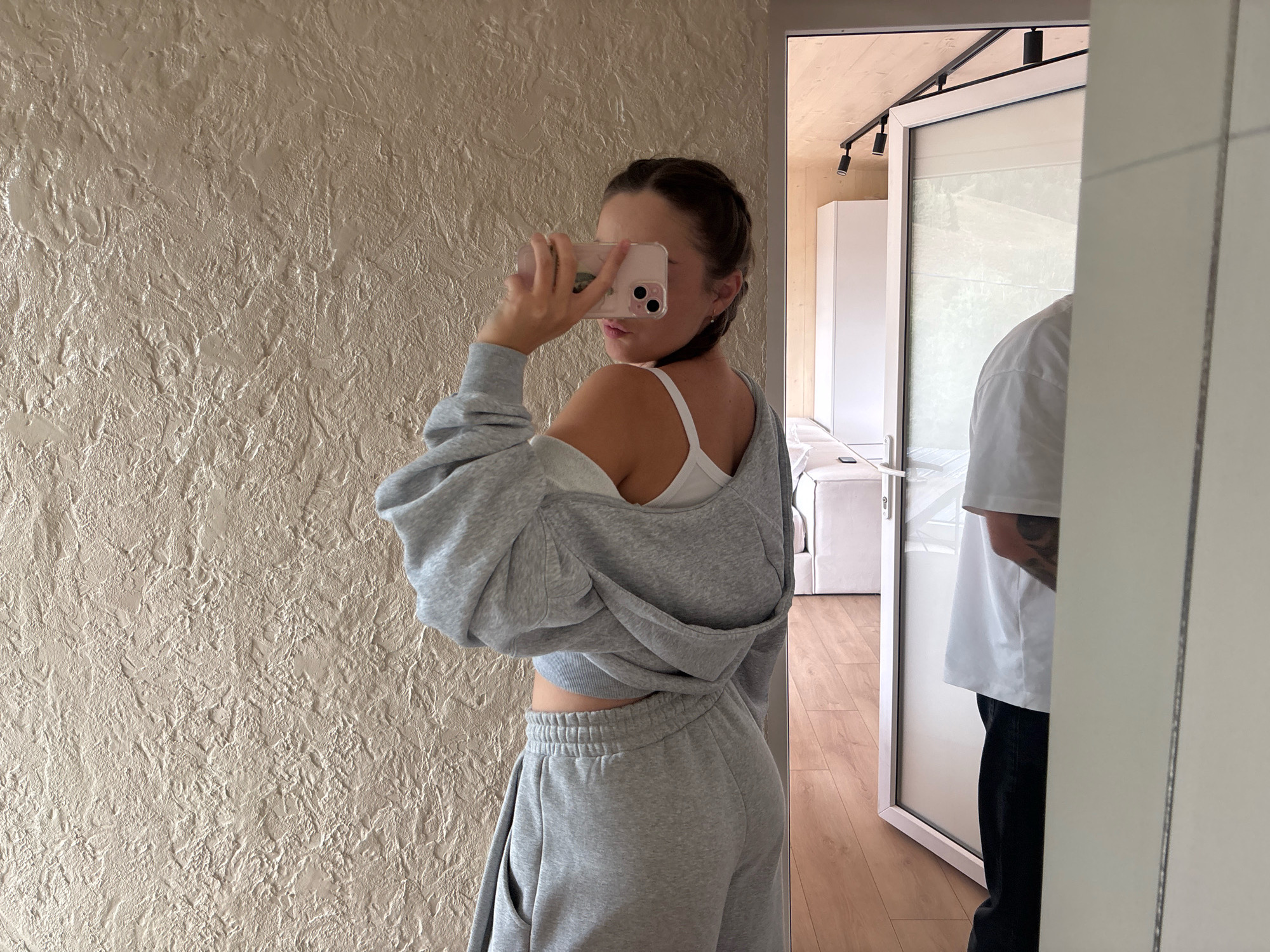 In Krasnyk, Ukraine, a young woman is captured in a mirror selfie, her back to the camera. She wears a grey loungewear set, with a cropped hoodie casually draped off one shoulder, revealing a white tank top. Her hair is styled in a braid, and she holds a pink phone, obscuring her face. To her right, a man is partially visible in the reflection, wearing a white t-shirt and dark pants, and appears to have tattoos. The background shows a textured beige wall, a contemporary interior with wooden accents, and a glimpse of furniture, suggesting a modern home or apartment.