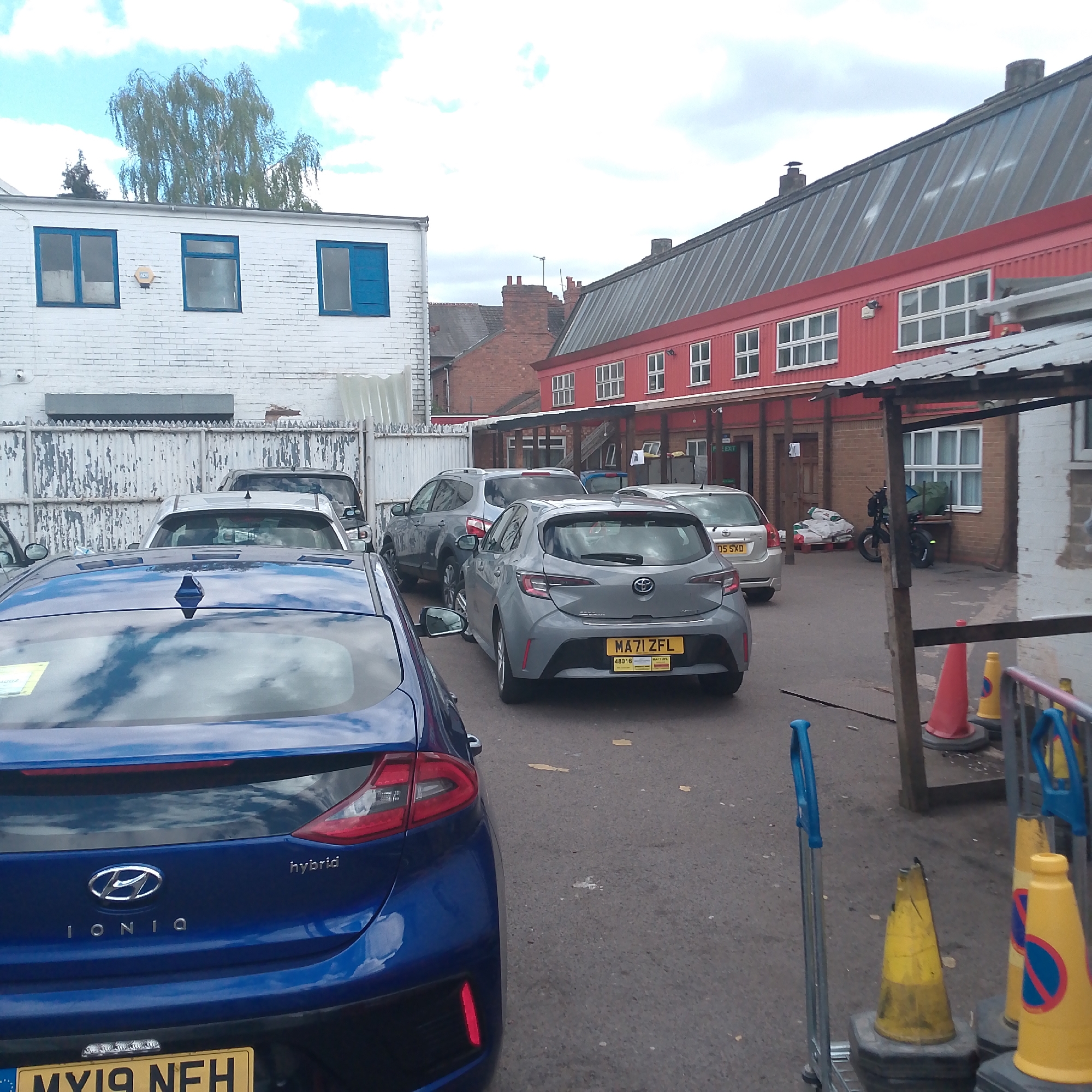 This daytime image, set in Coventry, United Kingdom, depicts a functional parking area or yard under a cloudy but bright sky.
In the immediate foreground on the left, the rear of a blue Hyundai Ioniq Hybrid is visible, featuring "HYBRID" and "IONIQ" badging and a license plate partially readable as "MY19 NFH". To the right, a grey Toyota Corolla is parked, displaying the license plate "MA71 ZFL" and a small yellow label with "4B016". Further back, a silver car with the license plate "DS05 SXD" and a grey Nissan Qashqai are also parked.
On the left side of the scene, a white brick building with three blue-framed windows stands prominently. An orange alarm box is affixed to its facade. Below this building, a white wooden fence with weathered, peeling paint separates the building from the parking area. Above the fence, an air conditioning unit is visible. Behind the white building, a tall, weeping willow-like tree reaches towards the sky.
To the right, a long, red building with a distinctive red metal roof stretches across the midground. It features several white-framed windows and a covered walkway or awning running along its length. Outside this building, near a doorway, pallets stacked with white sacks are visible alongside a black bicycle or e-bike. A partially obscured "EXIT" sign can be seen above one door.
The right foreground contains several orange and yellow traffic cones, some bearing red circular "no waiting" or "no stopping" symbols. A blue-handled metal hand truck is also partially visible. The ground is asphalt. In the distance, behind the red building, brick buildings with chimneys are visible, suggesting an urban setting. The overall atmosphere is utilitarian, devoid of human activity or explicit emotions.
