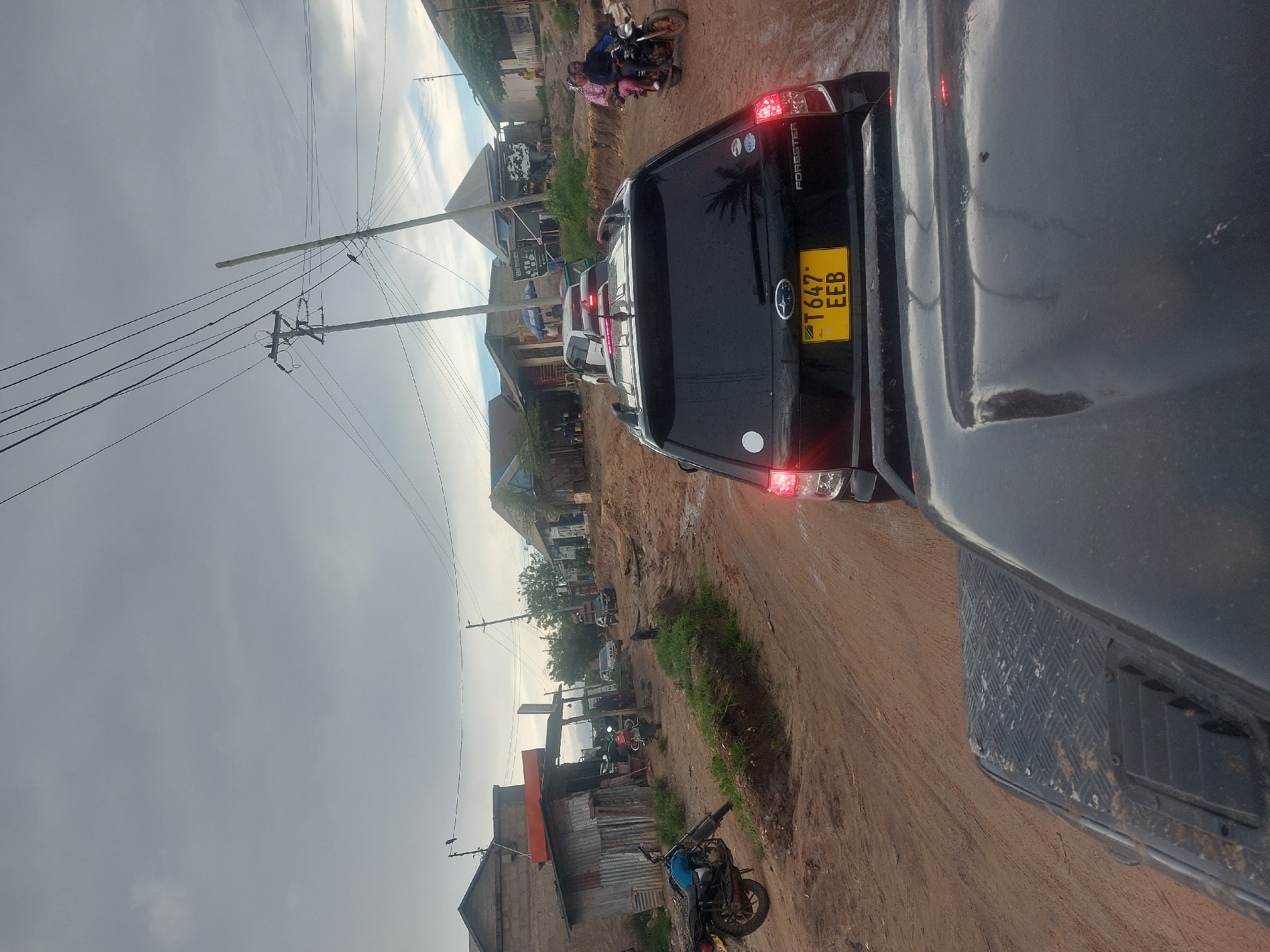 The image shows a muddy street scene in Dar es-Salaam, Tanzania. A black Subaru Forester, registration number T 647 EEB, is in the foreground, partially obstructing the view. The street is unpaved and muddy, with buildings lining both sides. Several motorcycles are visible, one with two people on it. Power lines stretch across the sky, which is overcast. Other vehicles are visible in the distance.