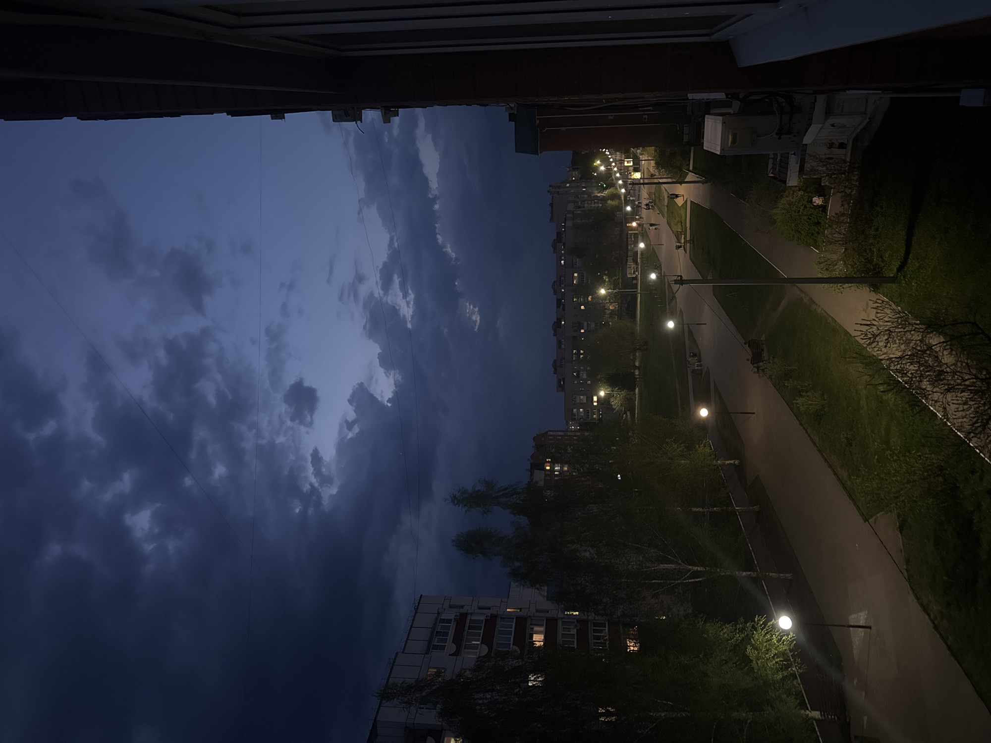 The image shows a nighttime view from a high vantage point, likely an apartment window, in Chaikovsky, Russia. The sky is dominated by dark, ominous clouds. Below, a residential area is visible with apartment buildings illuminated by internal lights. A pedestrian walkway runs through a green space, lit by streetlights. Several people can be seen walking along the path. The environment suggests a typical urban residential area at twilight or early night.