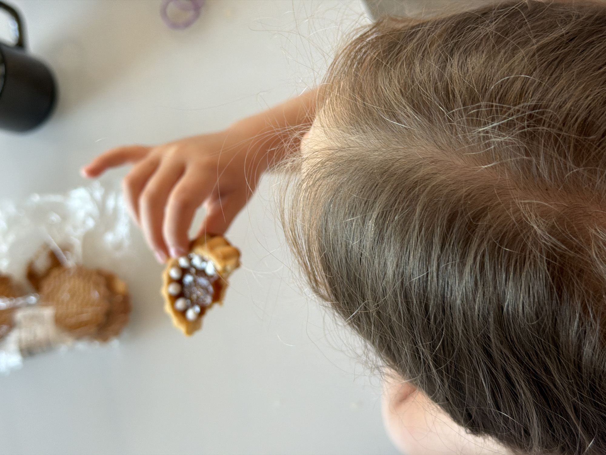 A child is reaching for a tart cookie filled with caramel and white sprinkles. The child's light brown hair is visible in the foreground. The cookie is held in the child's left hand. Other cookies in a wrapper are visible on the white surface. A black mug and a purple hair tie are also on the surface. The location is unknown.