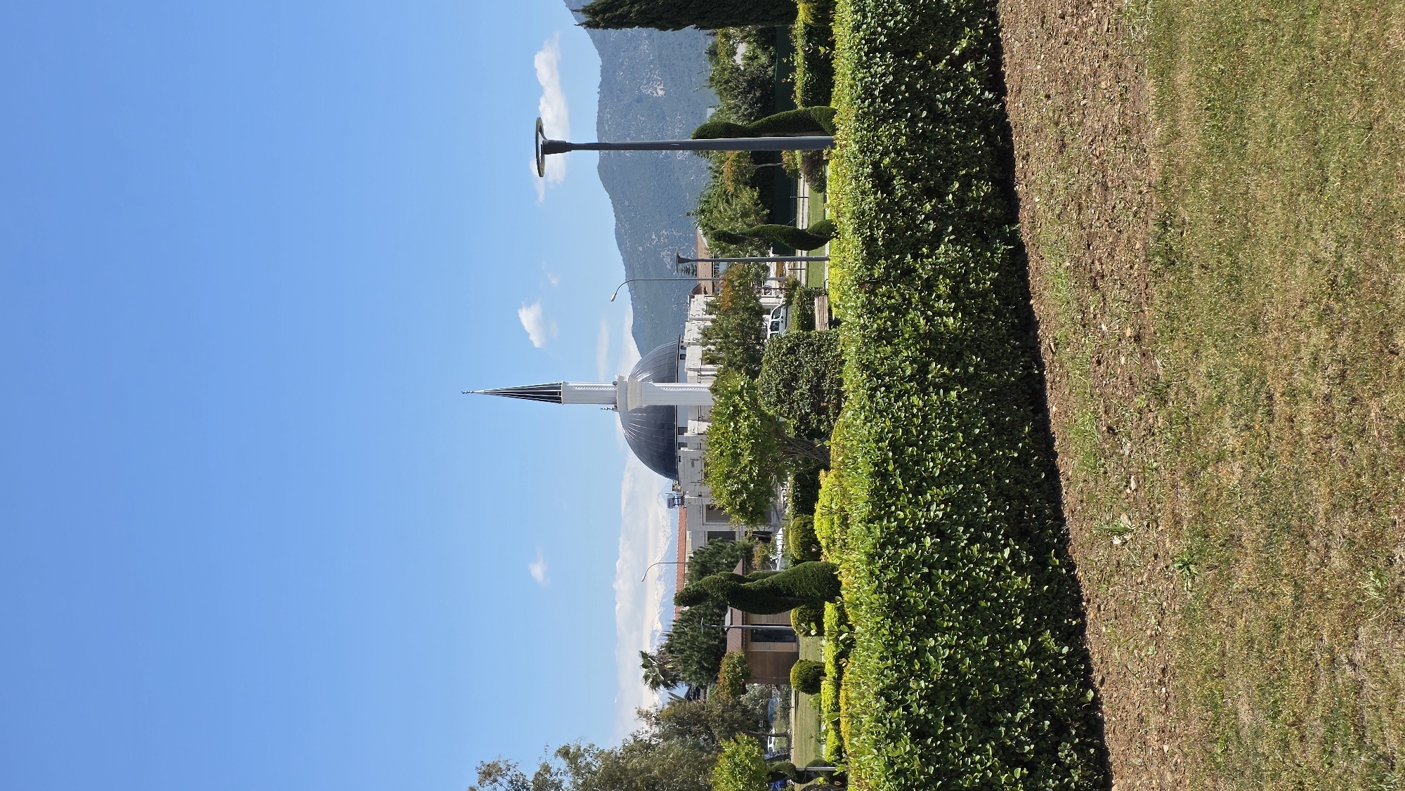 The image depicts a park scene in Fethiye, Turkey, featuring the Fethiye Merkez (Central) Mosque. The mosque is visible with its characteristic white minaret and dark dome, set against a backdrop of mountains under a clear blue sky with a few clouds. Streetlights are visible in the distance. The foreground shows manicured greenery, including a neatly trimmed hedge and a patch of dry, somewhat patchy grass. The image is rotated 90 degrees counterclockwise. There are no people visible in the image.