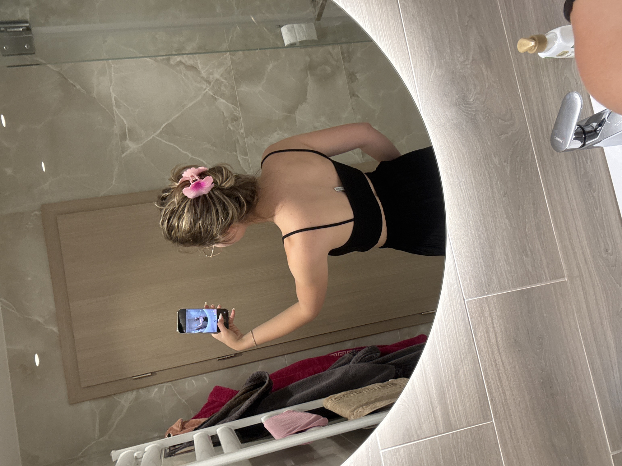 A young female is depicted from the back, holding a smartphone in her right hand to capture a mirror selfie within a modern bathroom. Her blonde hair is styled in a messy bun, secured by a prominent pink hibiscus-shaped hair clip. She wears a black spaghetti-strap crop top, revealing a white tag or label on the inner back, and black bottoms. A thin gold-colored bracelet adorns her right wrist, and eyeglasses are visible on her right ear.
The bathroom features a large, circular mirror with an illuminated edge. The walls are composed of light grey marble-patterned tiles on the upper section and lighter, horizontally-oriented wood-grain patterned rectangular tiles below. Reflected on the left side of the mirror is a white ladder-style towel warmer holding multiple towels or garments in dark grey, red, pink, and beige. A light-colored wooden door with a vertical handle is also reflected. To the right of the subject, a clear glass shower partition with a metal hinge is visible, behind which a roll of white toilet paper is mounted on a holder. The foreground shows a white sink basin equipped with a chrome single-handle faucet, and a white bottle with a gold pump dispenser (likely soap or lotion) rests beside it. The precise location within Sofia, Bulgaria, cannot be determined from the visual information available in the image.
