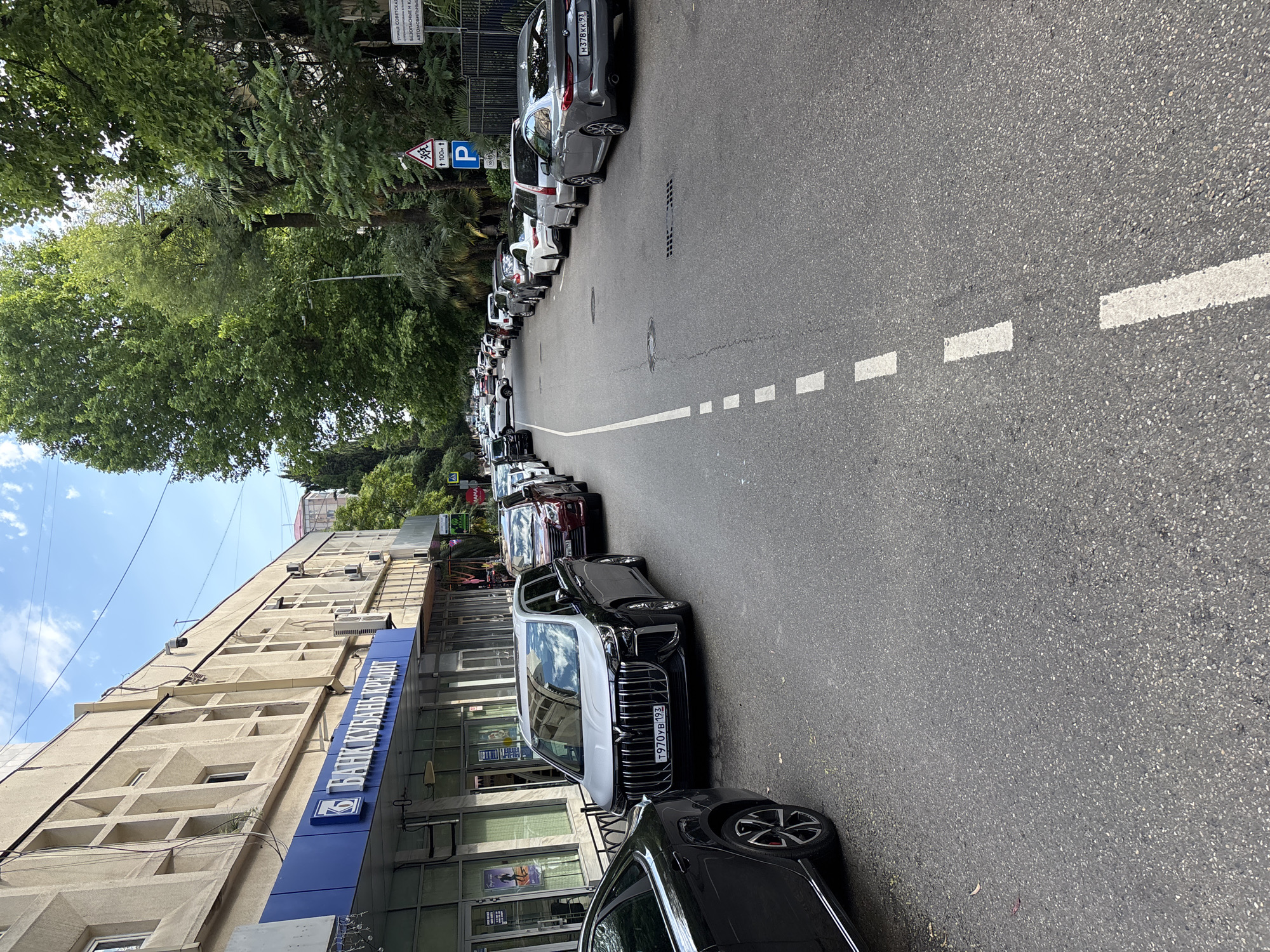 This media file shows a street scene in Sochi, Russia, with a row of parked cars lining the side of the road. The perspective is from the sidewalk, looking down the street. A long, light-colored building with many windows runs along the left side of the image. A blue sign on the building reads "БАНК КУБАНЬ КРЕДИТ". Several cars are visible, including a white BMW with the license plate "T970УВ 193" and a grey BMW with the license plate "M378KK 93". Traffic signs are also visible, including a parking sign with a blue 'P' and a pedestrian crossing sign. Trees and greenery are present, suggesting an outdoor setting on a bright, possibly sunny day. The scene appears to be a typical urban environment with shops and residential buildings. No people are prominently visible in the image. The presence of parked cars suggests it is daytime. The overall mood is neutral, simply documenting the streetscape.