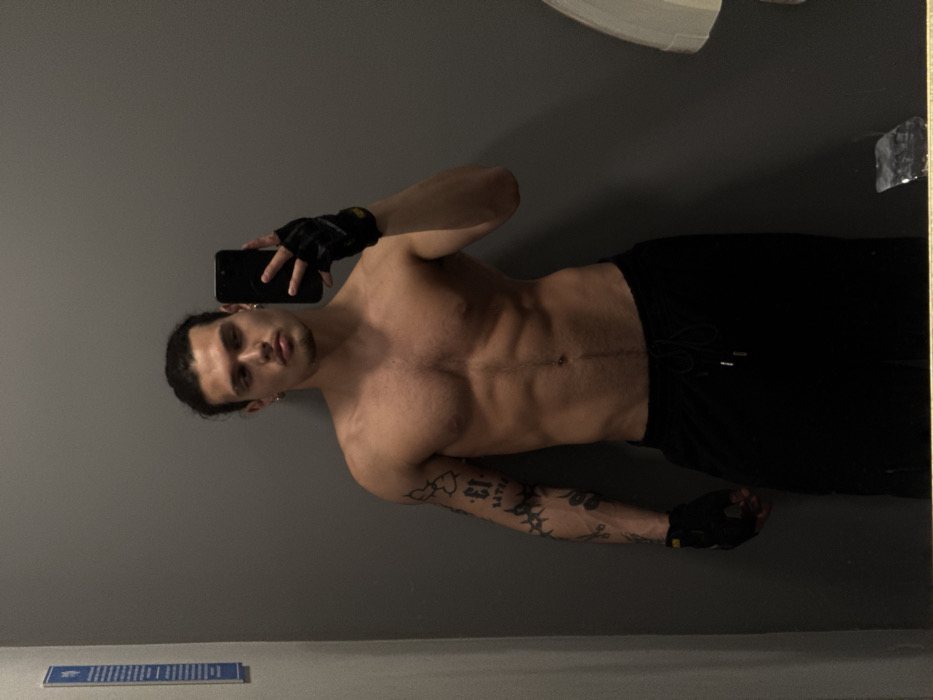 An indoor mirror selfie depicts a shirtless, muscular young adult male located in Sofia, Bulgaria. The subject stands with his torso angled slightly, holding a black smartphone in his gloved right hand to capture his reflection. He wears black fingerless athletic gloves on both hands. His dark hair is pulled back. He presents a defined physique with visible chest hair, a navel piercing, and distinct abdominal muscles. His left arm is heavily tattooed with various designs, including the number "13" and abstract patterns. He is clad in black drawstring athletic pants. The background behind the subject's reflection is a uniform gray wall. A blue and white rectangular sign is visible mounted on the wall to the far left. A partial reflection of a white, possibly ceramic fixture, is discernible in the mirror on the far right edge, suggesting a restroom or changing room environment.