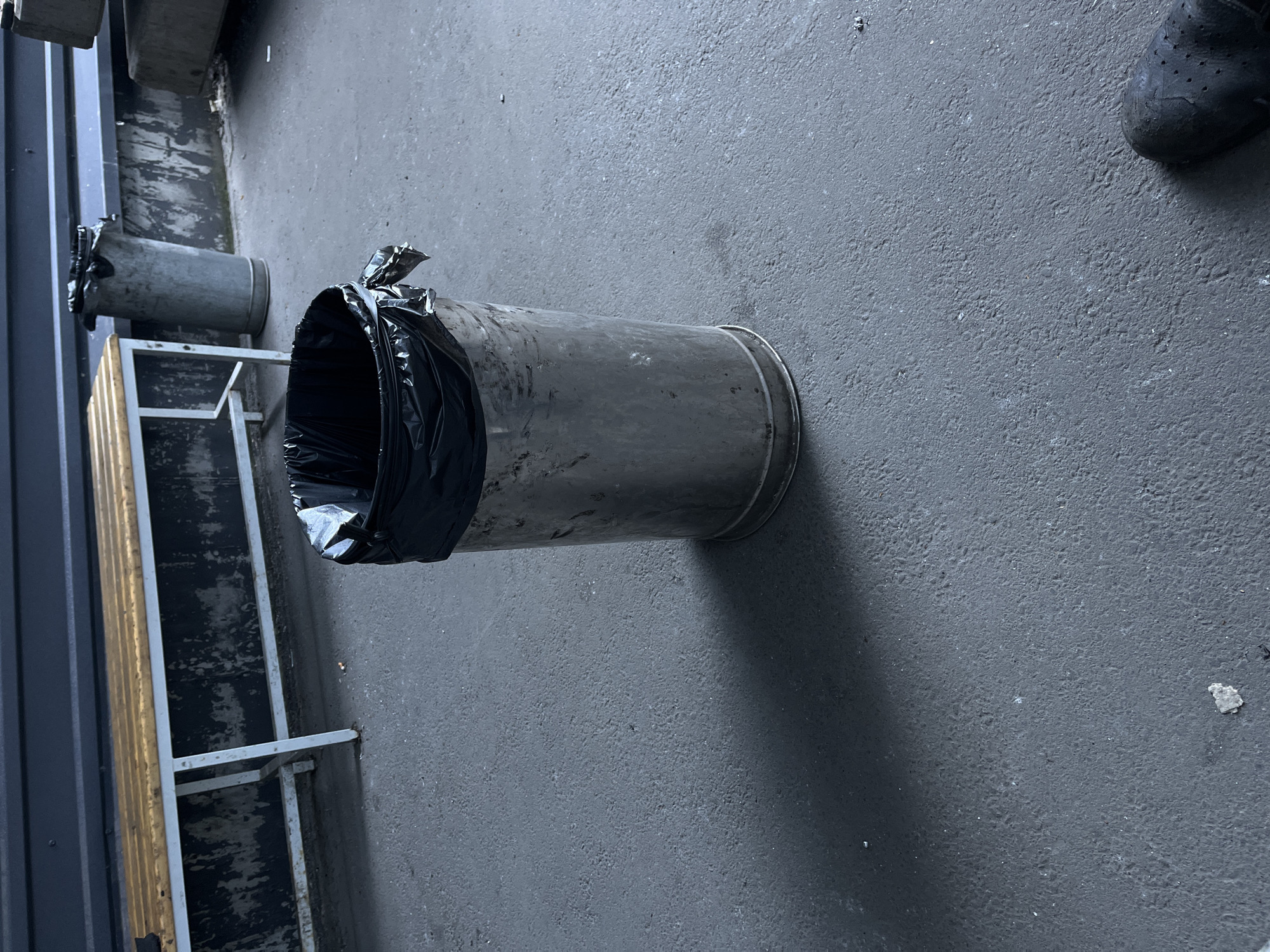 The image depicts an outdoor scene, likely in an urban setting. In the foreground, a cylindrical metal trash can is present, containing a black trash bag that is visibly overflowing. The trash can is positioned on a grey asphalt surface. A shadow is cast by the trash can to its left, indicating an overhead light source. In the background, a wooden bench with a metal frame is visible, and behind that, a dark, textured wall. Further in the background, another, larger metal trash receptacle can be seen. The overall impression is one of an outdoor public space, possibly a waiting area or a rest stop. The location within Kyiv, Ukraine is not specified in the image.