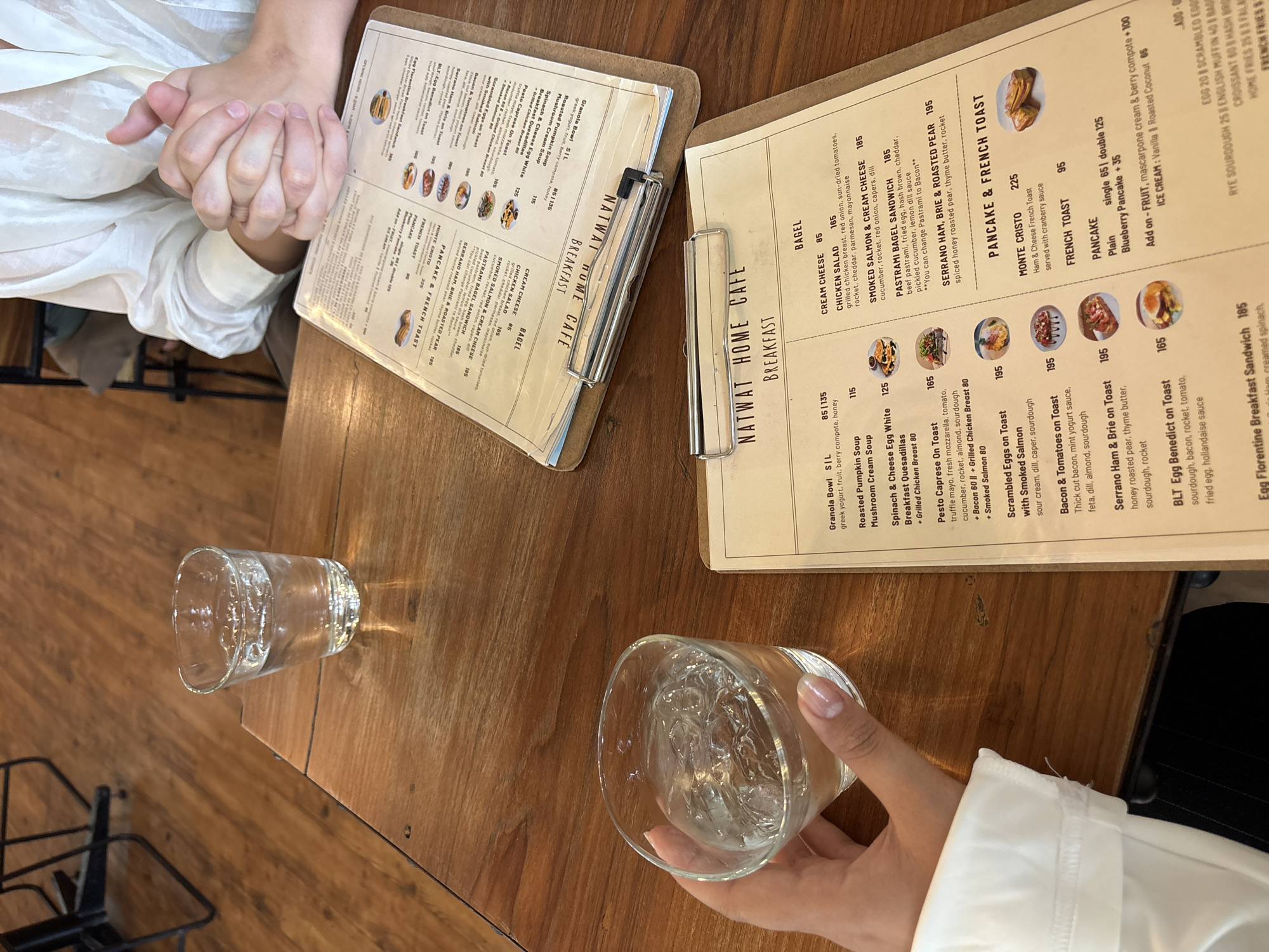 The image depicts a scene at a cafe, likely in Chiang Mai City Municipality, Thailand, given the menu items. The focus is on a wooden table with two menus and two glasses of water. In the foreground, a person's hands are clasped together, wearing a light-colored shirt, and they are positioned over one of the menus. To the left, another hand is holding a glass of water with ice. In the background, another person's hands are visible, also clasped. The menus are for "NATWAT HOME CAFE" and list breakfast items such as "Granola Bowl," "Roasted Pumpkin Soup," "Spinach & Cheese Egg White Breakfast Quesadillas," "Pesto Caprese On Toast," "Scrambled Eggs on Toast with Smoked Salmon," "Bacon & Tomatoes on Toast," "Serrano Ham & Brie on Toast," "BLT Egg Benedict on Toast," and "Egg Florentine Breakfast Sandwich." The "BAGEL" section includes "Cream Cheese," "Chicken Salad," and "Smoked Salmon & Cream Cheese." The "PANCAKE & FRENCH TOAST" section lists "Monte Cristo" and "French Toast." The environment appears to be a cafe with wooden furniture and flooring.