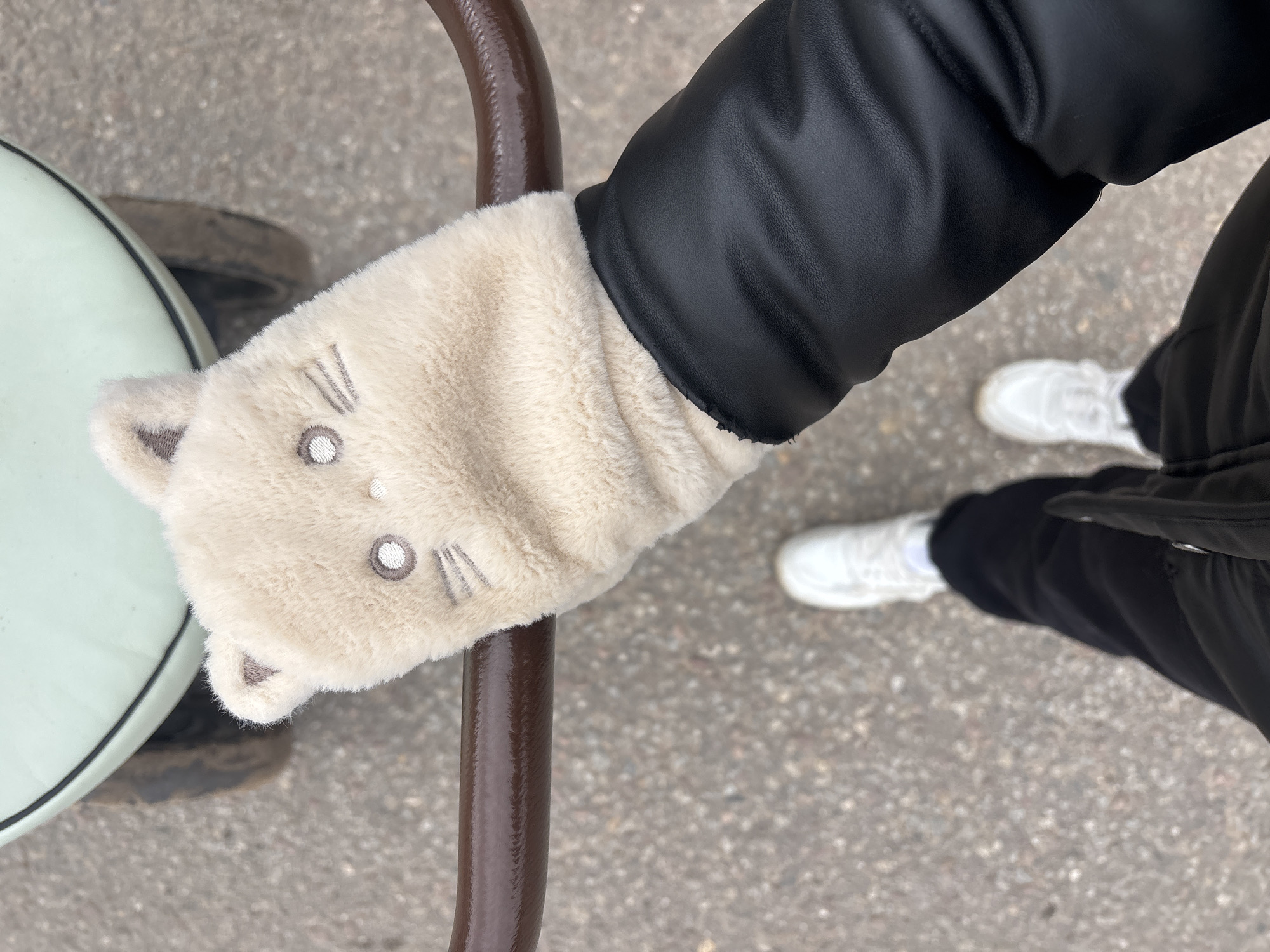 A person in a black jacket and black pants is holding onto a metal bar. They are wearing a light-colored mitten with cat-like features on their right hand. White sneakers are visible on the person's feet. The location is a paved surface, likely a street or walkway. The location is within the city of Раздолье, Russia.