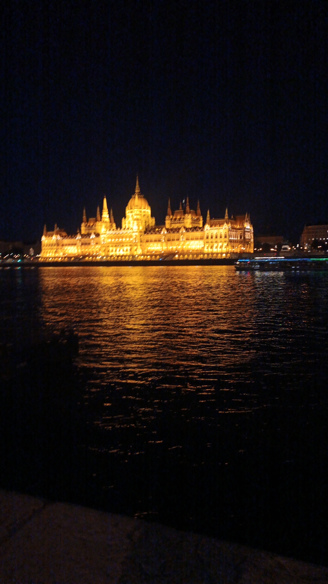 The image captures the Hungarian Parliament Building in Budapest, Hungary, illuminated at night. The building is reflected in the Danube River, with ripples on the water surface. A tour boat with lights is on the river. The foreground is dark, obscuring details of the riverbank. The sky is dark.