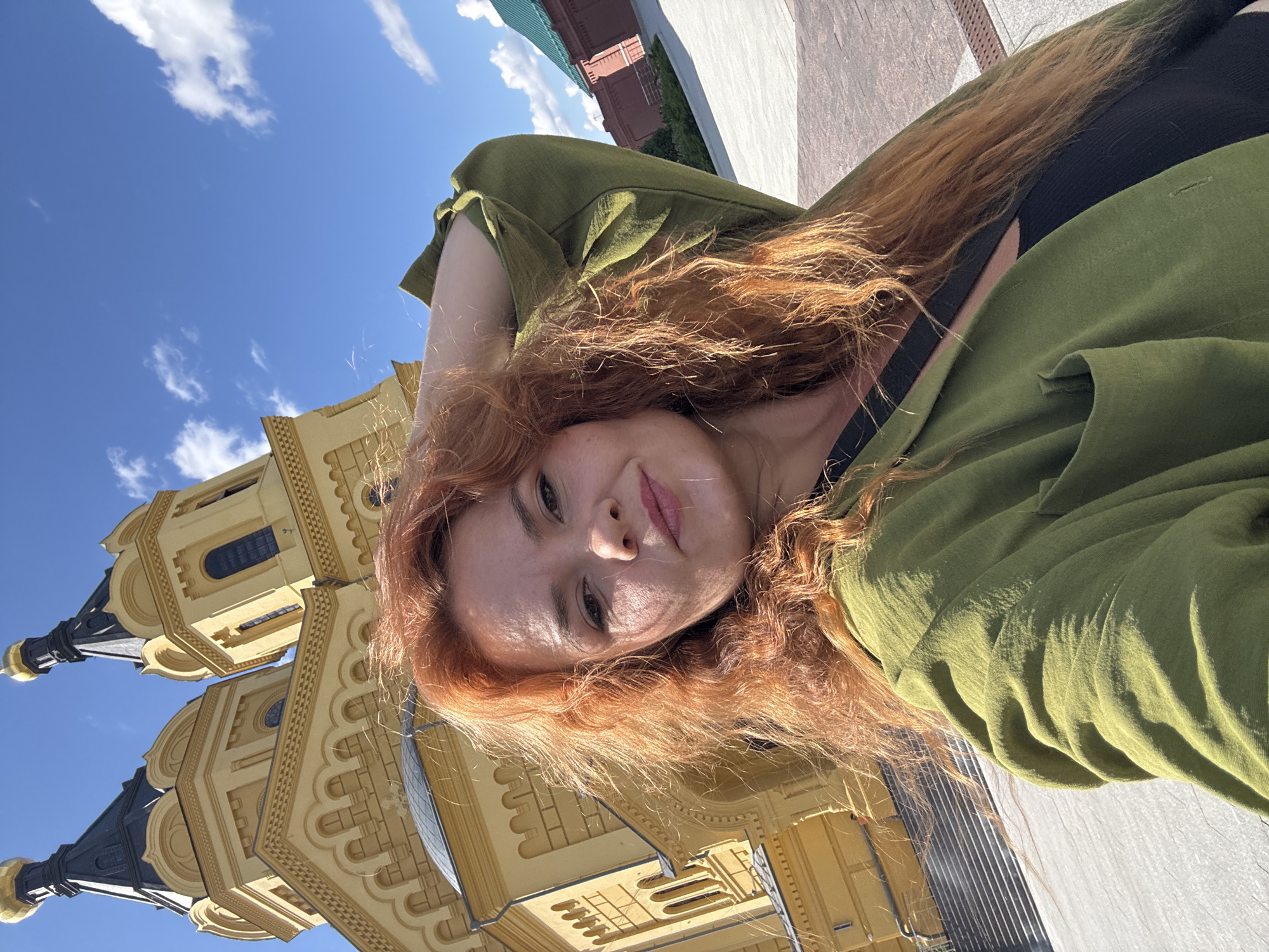 The image shows a woman with long, wavy red hair taking a selfie outdoors. She is wearing a green jacket over a black top and has her left arm raised, with her hand resting on her head. Behind her, a large, yellow building with multiple domes and architectural details is visible, under a bright blue sky with scattered white clouds. This building is identifiable as the Alexander Nevsky Cathedral in Nizhny Novgorod, Russia, a prominent landmark in the city. The area around the cathedral appears to be a paved courtyard or public space. The lighting suggests it is daytime, possibly late morning or afternoon, with sunlight casting shadows and highlighting the woman's hair and skin.