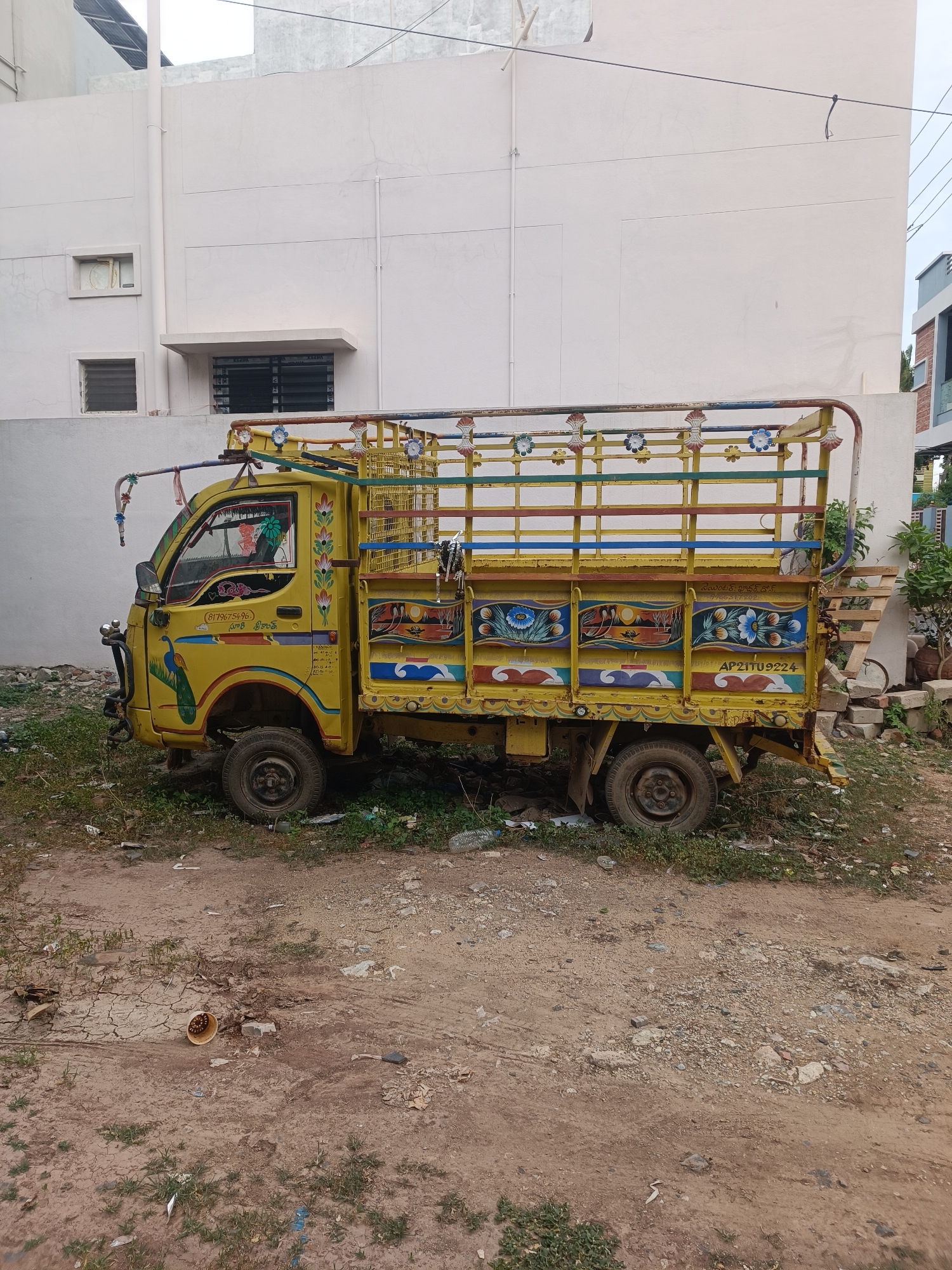 The image depicts a yellow, decorated, three-wheeled truck parked on a dirt lot with sparse vegetation. The truck features colorful floral and peacock motifs painted on its body, with a yellow cage-like structure on the back. A license plate reading "AP21TU9224" is visible on the rear of the truck. In the background, a light-colored building with windows and a pipe extending vertically along its exterior is present. The ground is covered with dirt, scattered debris, and some patchy grass. To the right of the truck, a small pile of bricks and some potted plants are visible. The sky is overcast. The scene appears to be in an urban or semi-urban area, likely in Bethamcherla, India, given the license plate format. There are no people or animals visible in the image.