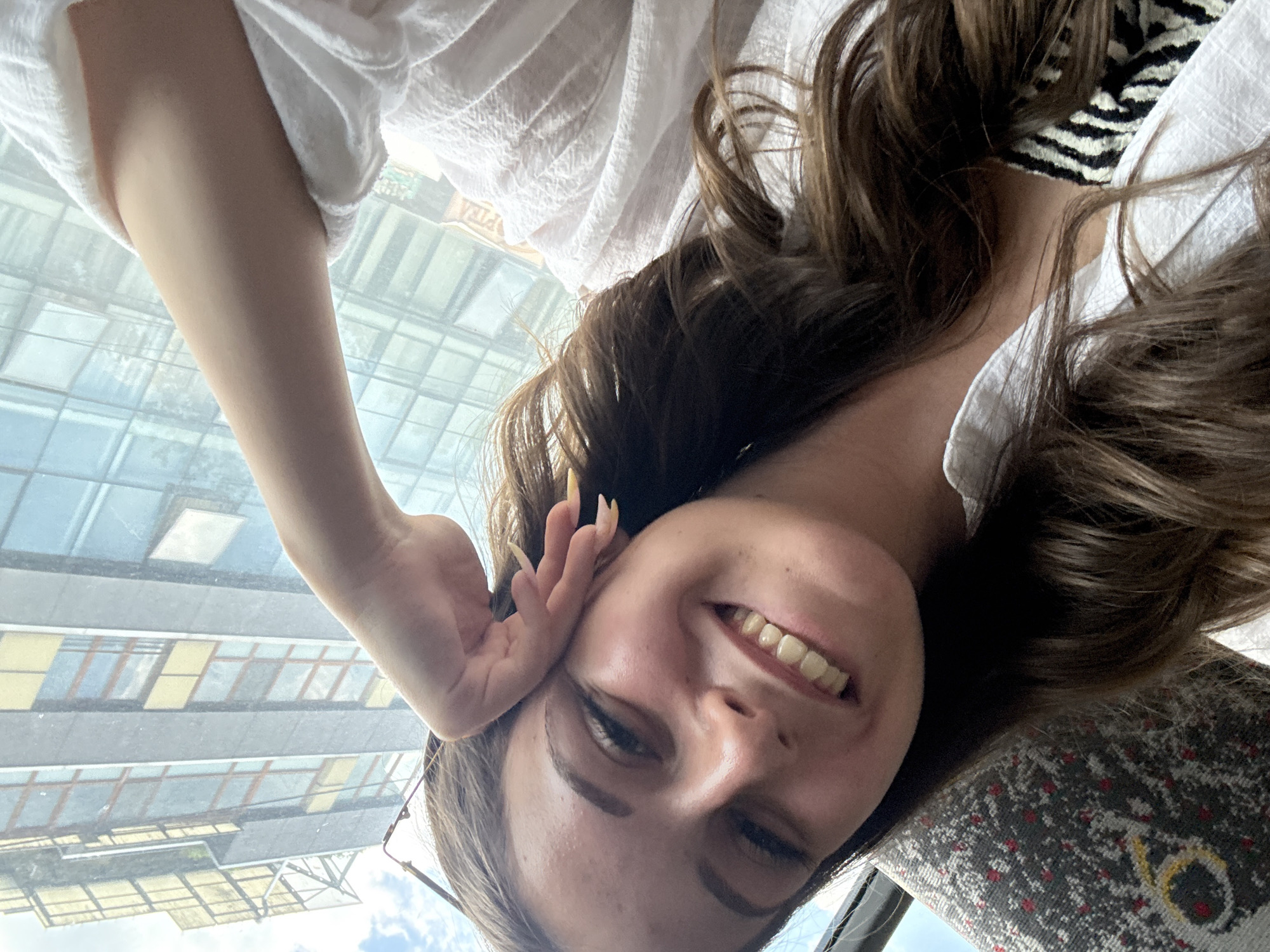 The image shows a young woman taking a selfie, with the camera rotated 90 degrees clockwise. She has long, dark brown hair, and is wearing a white shirt with a textured fabric. A black and white striped pattern is visible on the collar of her shirt. Her left hand is touching her ear, and she is smiling, showing her teeth. Her fingernails are long and painted. A pair of glasses is visible on her face.
The background appears to be a window overlooking a modern building with many windows and balconies. The building's architecture suggests an urban environment. The lighting suggests it is daytime, possibly with bright sunlight. The texture of a patterned seat is visible in the lower right corner, suggesting she may be seated on public transportation.
The overall mood of the image is cheerful and relaxed. No visible text is present in the image. Given the location context, the scene is likely in Burgas, Bulgaria.