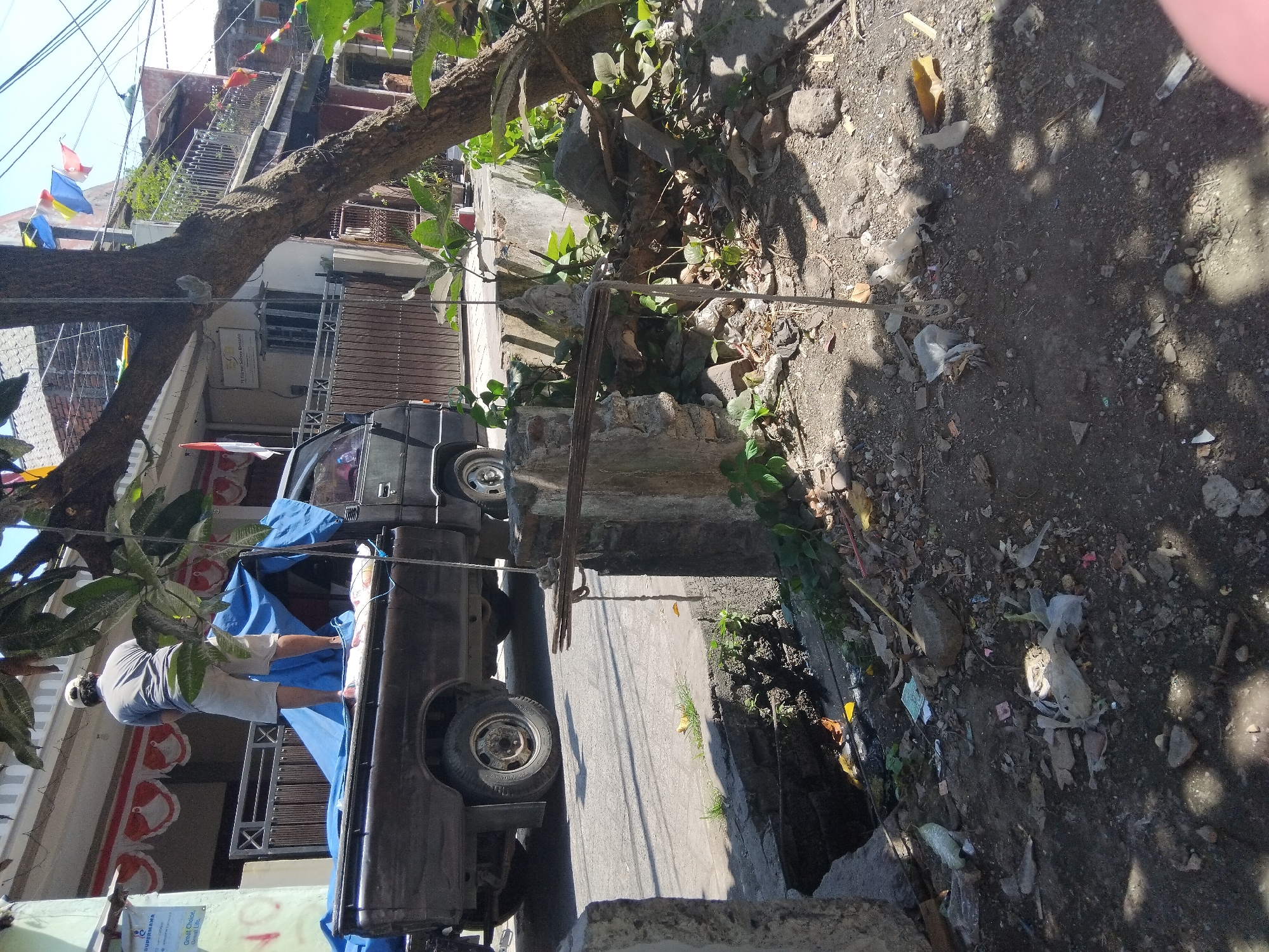 A man wearing a white cap and a gray t-shirt is standing on the back of a dark-colored pickup truck, which is covered with a blue tarp. He is wearing light-colored shorts and appears to be urinating. The truck is parked on the side of a street in Berbek, Indonesia. The street is lined with buildings, some of which have flags or banners displayed. There is a tree with a thick trunk and branches in the foreground, casting shadows on the ground. The ground is covered with dirt, rocks, and debris, including plastic bags and other trash. A concrete structure with some vegetation growing on it is visible to the right of the truck. A drainage ditch runs along the side of the street, also containing debris. The overall scene suggests a somewhat unkempt urban environment.