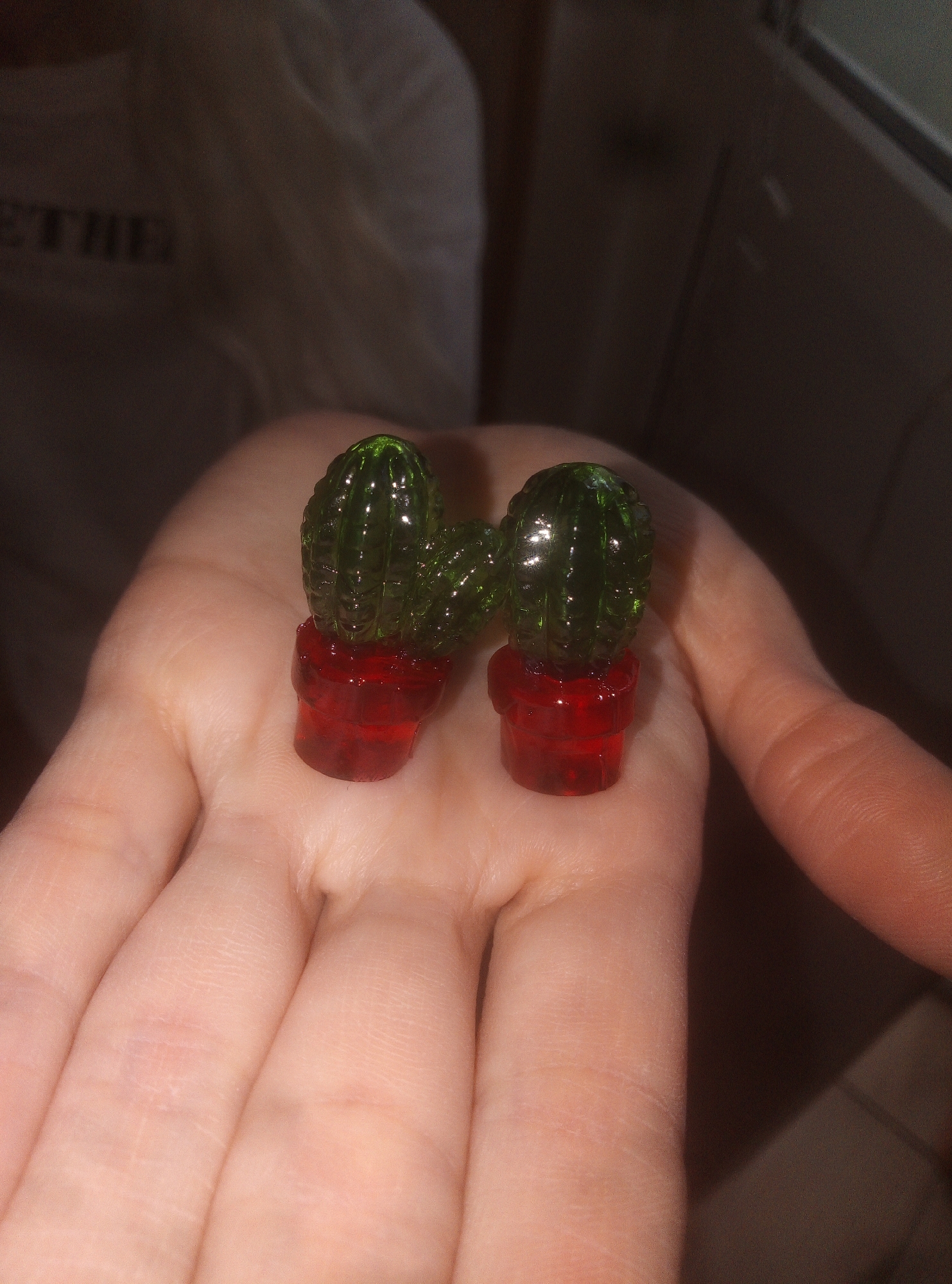 The image presents a close-up view of a person's open hand, holding two small, green, translucent gummy candies. Each candy is intricately shaped like a cactus and is placed within a small, translucent red base designed to resemble a plant pot. The candies exhibit a glossy, slightly bumpy surface, reflecting light and highlighting their gelatinous texture.
The hand, which is the primary subject, shows visible skin texture, creases, and lines on the palm and fingers, suggesting a person with light skin. The background is softly blurred, indicating a shallow depth of field. In the upper left background, part of another person's torso or clothing is visible, and on their light-colored fabric, the partial text "ETHE" can be discerned. The setting appears to be indoors, with ambient or artificial lighting that illuminates the candies from the side, emphasizing their translucency. No specific time of day, weather conditions, or strong emotions are evident.