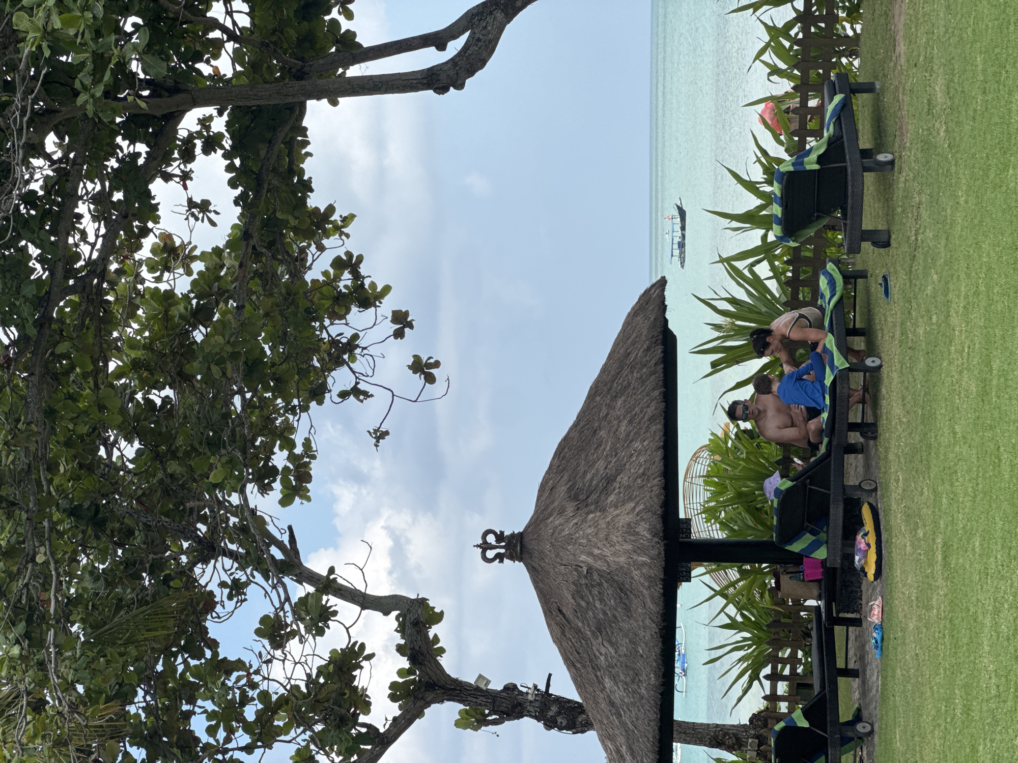 In Kuta Selatan, Indonesia, on a bright day, a family is relaxing under a thatched umbrella on a resort lawn. A man and a child are seated on lounge chairs, with a woman joining them. The chairs are arranged on a well-maintained grassy area with lush green plants and a wooden fence in the background. Beyond the fence, the turquoise ocean stretches to the horizon, with a small boat visible on the water. The scene evokes a sense of tranquility and vacation, as the family enjoys the tropical scenery.