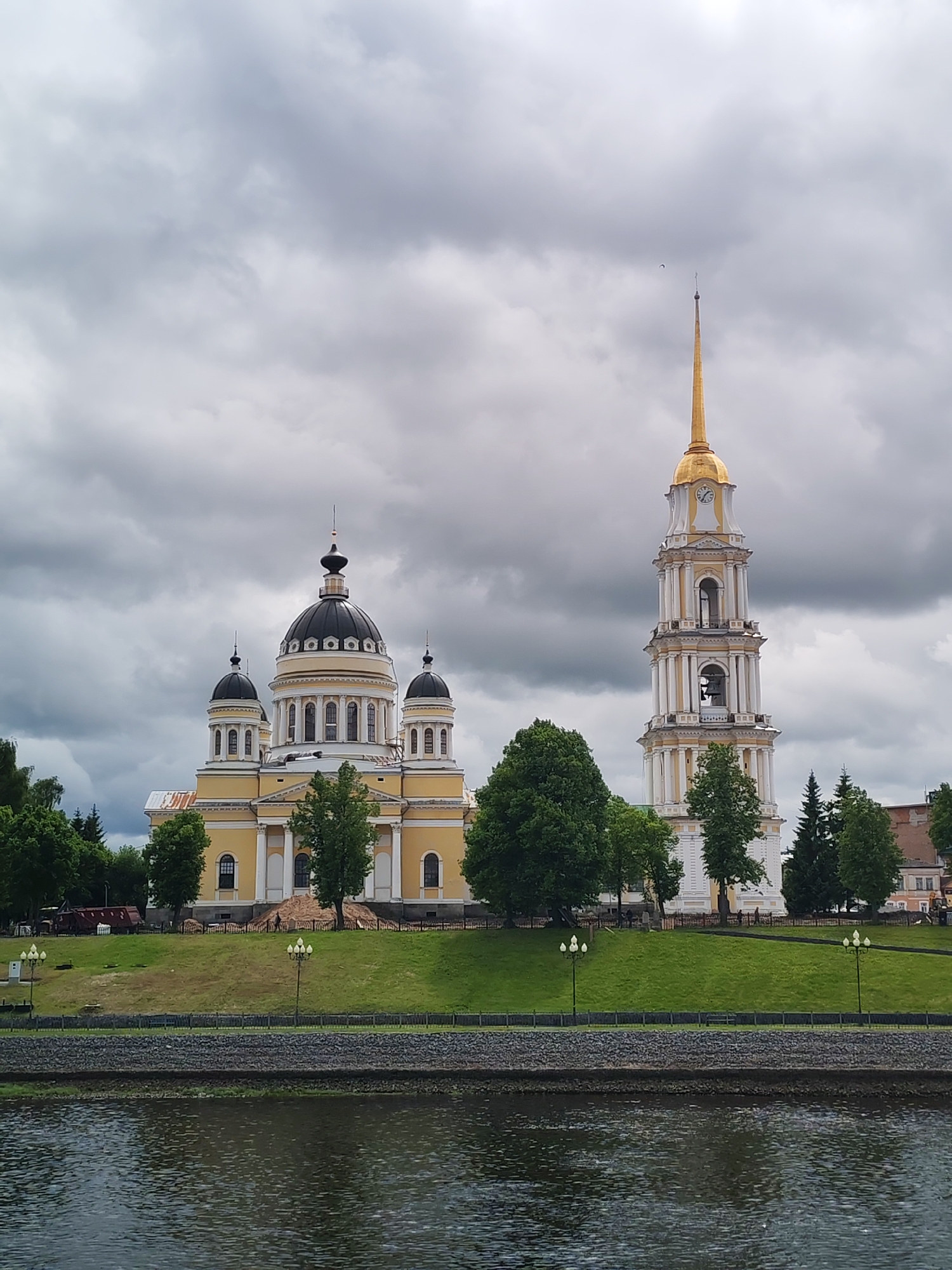 The image shows a view of the Transfiguration Cathedral and its bell tower in Rybinsk, Russia, from across the Volga River. The cathedral, painted in yellow and white, has a central black dome and smaller domes on the four corners. The bell tower is tall and white with a golden spire. The sky is overcast with heavy, gray clouds. The river is in the foreground, with a stone embankment. A grassy slope leads up to the cathedral and tower, dotted with trees and lampposts. Some human figures are visible on the slope.