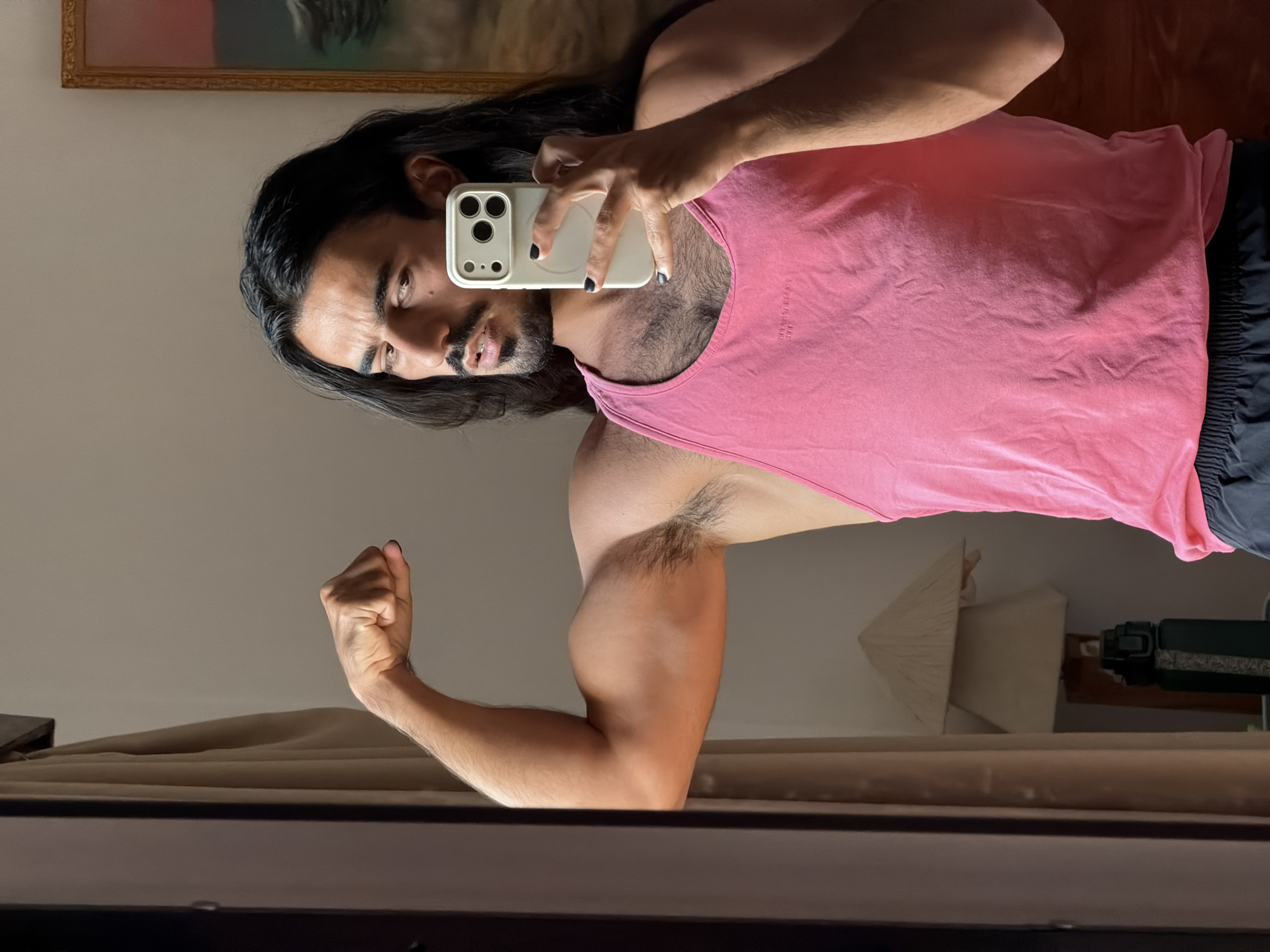A person with long, dark hair is taking a mirror selfie in what appears to be a bedroom. They are wearing a pink tank top and black shorts. The person is flexing their bicep, showcasing their arm muscles. In their right hand, they are holding a white smartphone with a triple-lens camera, which is reflecting in the mirror. The background shows a wall with a framed picture and some curtains. A lamp or decorative item is visible on a shelf behind them. The overall lighting suggests it might be daytime, possibly late afternoon given the warm tones. The scene has a casual and personal feel, typical of a private setting like a home. There is no visible text in the image, other than what might be on the phone screen or clothing, which is not legible.