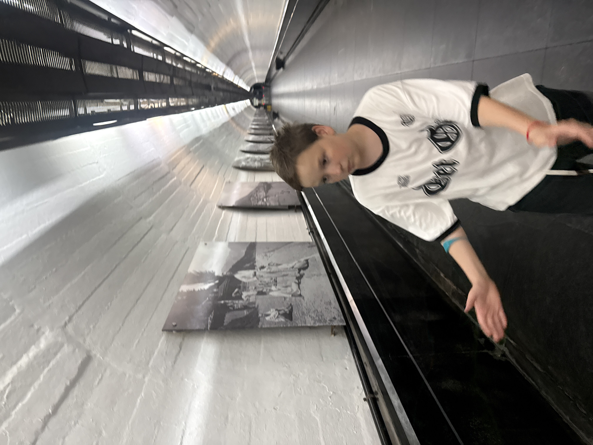 In the city of Yalta, Ukraine, a young boy with light brown hair is walking in a long, white-tiled tunnel. He is wearing a white t-shirt with black trim and a black logo, dark pants, and has a red bracelet on his right wrist and a blue band on his left arm. The boy is looking towards the right side of the frame and has his right arm extended outwards, palm open. On the left wall of the tunnel, there are several black and white photographs displayed, depicting groups of people in traditional clothing and mountainous scenery. The tunnel has a moving walkway on the right side of the frame. The ceiling of the tunnel is made of metal grating and lighting fixtures. The overall environment appears to be a public, possibly historical, passageway.