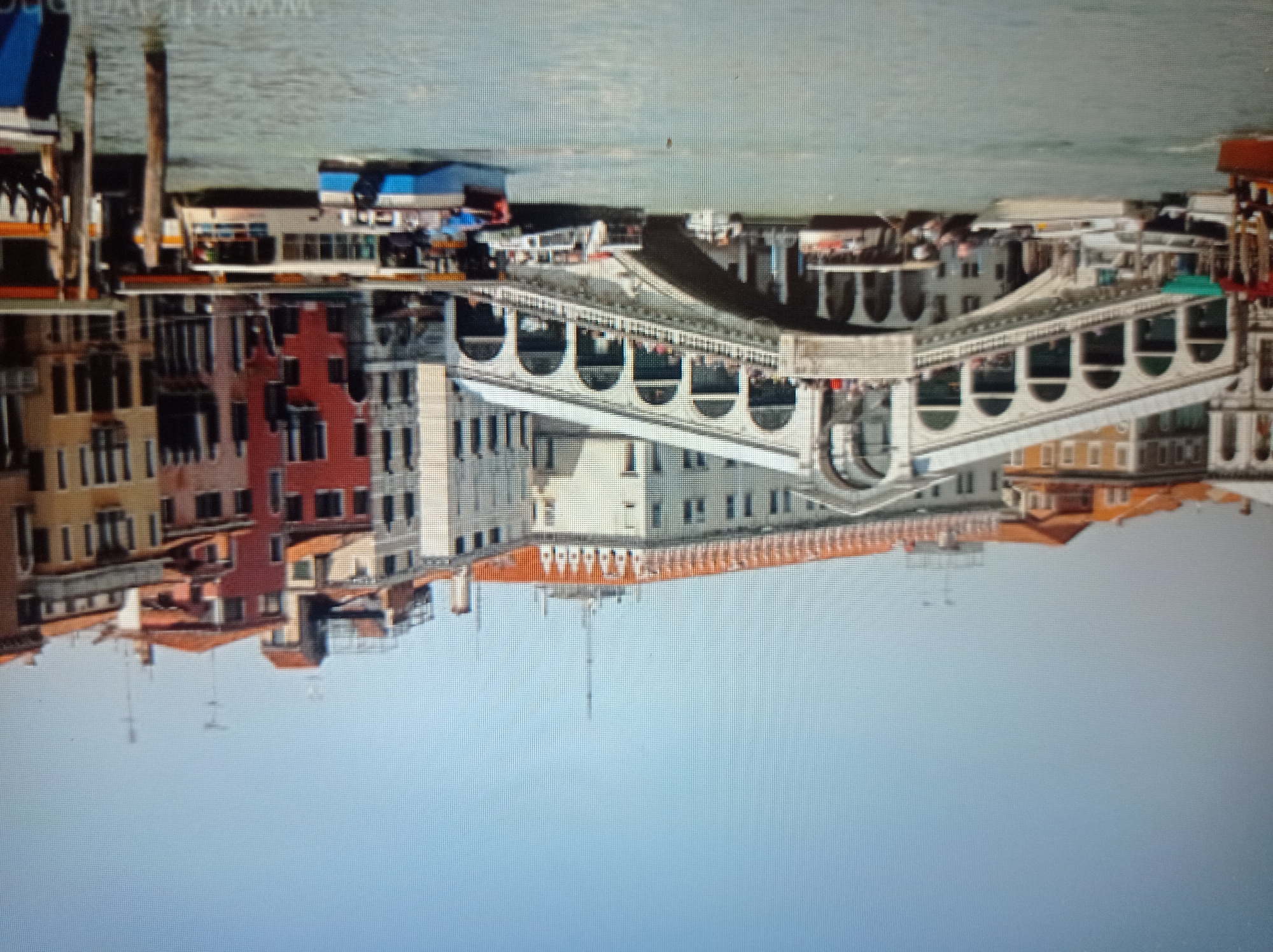 The image depicts the Rialto Bridge in Venice, Italy. The bridge is a stone arch bridge with shops built along its sides, spanning the Grand Canal. Several boats are visible in the canal, including passenger ferries and gondolas. Buildings with characteristic Venetian architecture, featuring colorful facades and arched windows, line the canal on either side. The sky is clear and blue, suggesting a sunny day. The scene is bustling with activity, with people visible on the bridge and on the boats, indicative of a popular tourist destination. The water of the Grand Canal is a murky green.