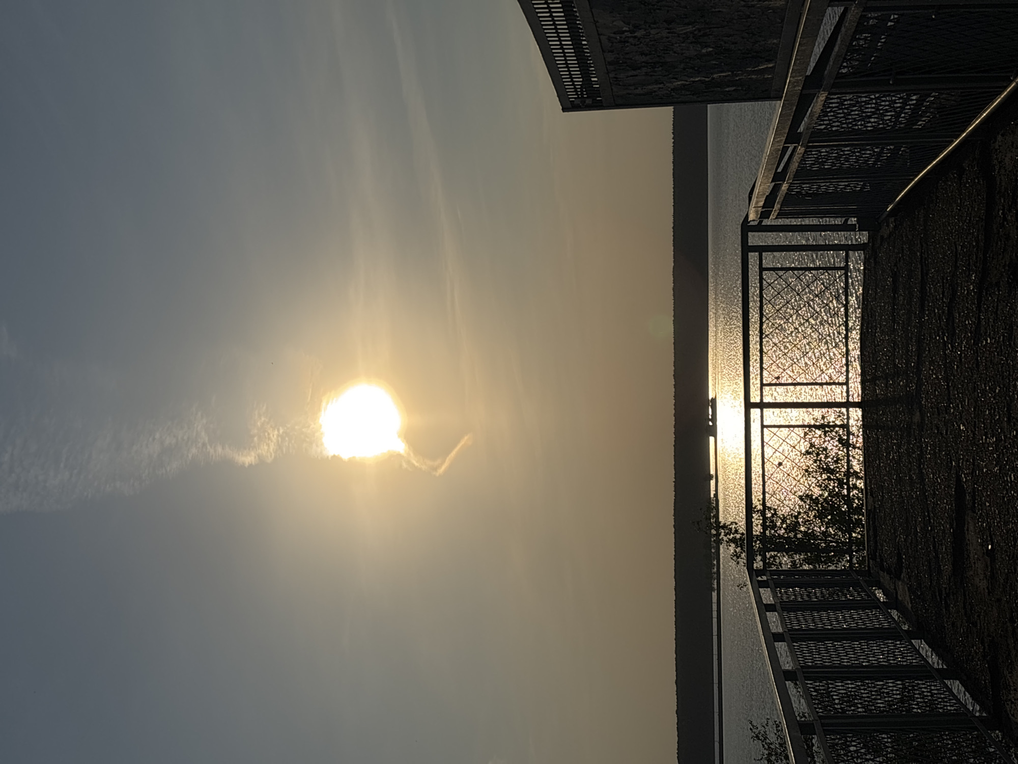 The image shows a sunset over the Kama River in Perm, Russia, viewed from a high point, possibly a bridge or elevated walkway, as indicated by the presence of a metal railing and a set of stairs. The sun is a bright, glowing orb obscured by a thin layer of clouds, casting a golden hue across the sky and reflecting in the water. The opposite bank of the river appears as a dark, indistinct line. A metal structure, possibly part of the elevated walkway, frames the right side of the image. Vegetation, possibly trees, is visible behind the metal structure. The lighting suggests it is either sunrise or sunset.