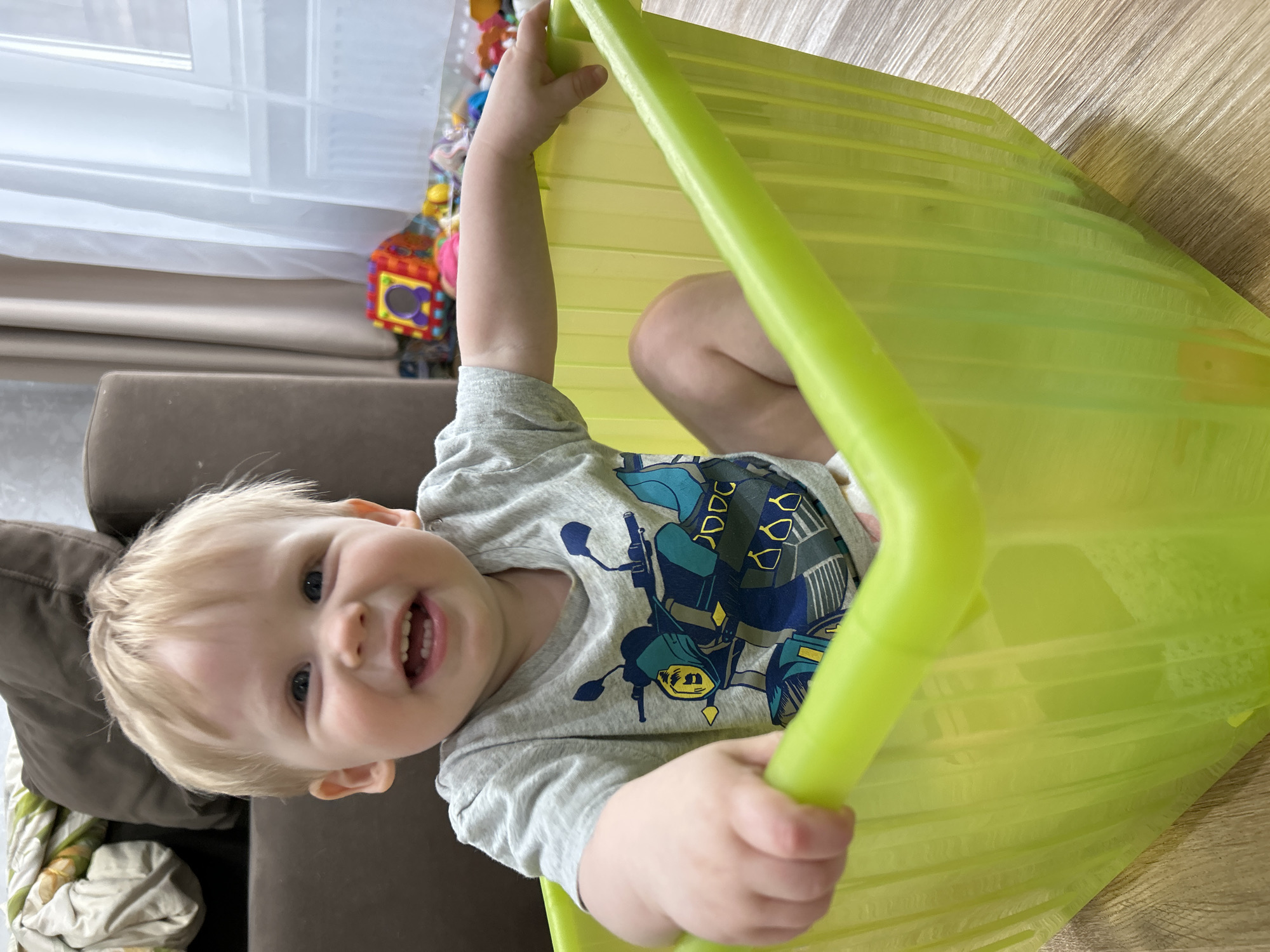 A young child with blonde hair and blue eyes sits inside a green plastic child's slide, indoors. The child wears a gray t-shirt with a motorcycle design. Part of a brown couch is visible to the left, and children's toys are in the background, near a window. The floor is light brown. The location is Chaikovsky, Russia.