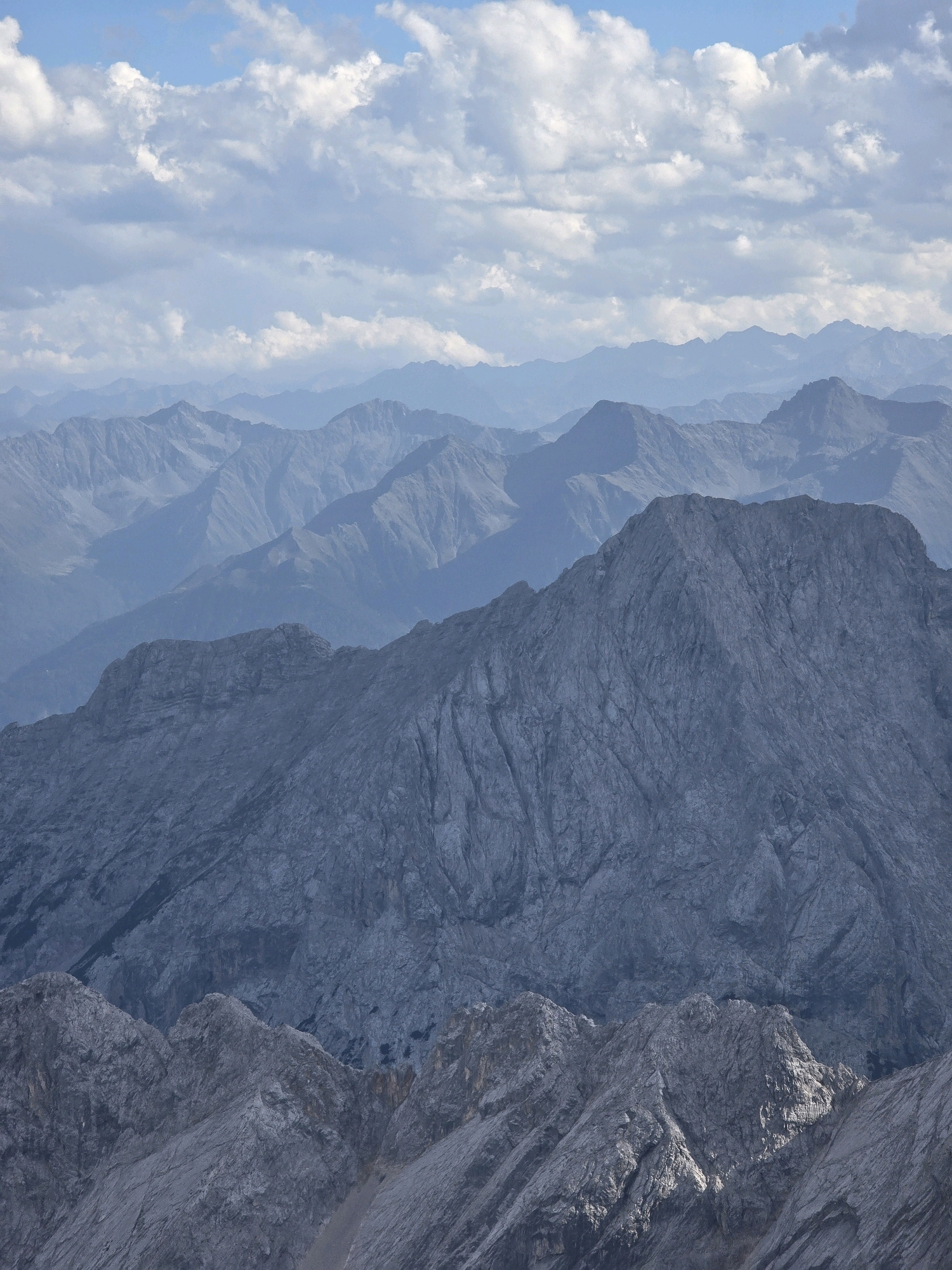 The image depicts a vast mountain range, likely the Alps, viewed from a high vantage point. The foreground is dominated by rugged, grey mountain peaks with sharp ridges and textured surfaces, suggesting rocky, uneven terrain. These prominent peaks recede into layers of more distant mountains, each appearing progressively hazier and bluer due to atmospheric perspective, creating a sense of immense depth. The sky above is a soft blue, dotted with scattered white, fluffy clouds, some of which are illuminated by sunlight, casting subtle shadows. The overall impression is one of grand, natural landscape, characterized by dramatic geological formations and a serene, expansive sky. This scene is representative of the mountainous environment surrounding Grainau, Germany, a town nestled in the Bavarian Alps known for its dramatic peaks and hiking opportunities. There are no people or man-made structures visible in the image, emphasizing the wild and natural character of the landscape.