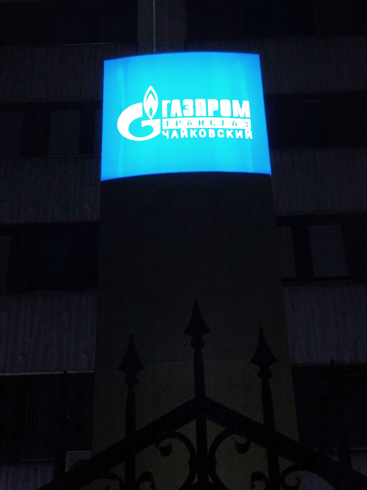 A lit, blue Gazprom sign is attached to a building. The sign includes the Gazprom logo, the name "TRANSGAZ," and the city name "CHAIKOVSKY" in Russian. A dark, wrought-iron fence is visible in the foreground. The background is a building with dark windows. The scene is likely at night in the city of Chaikovsky, Russia.