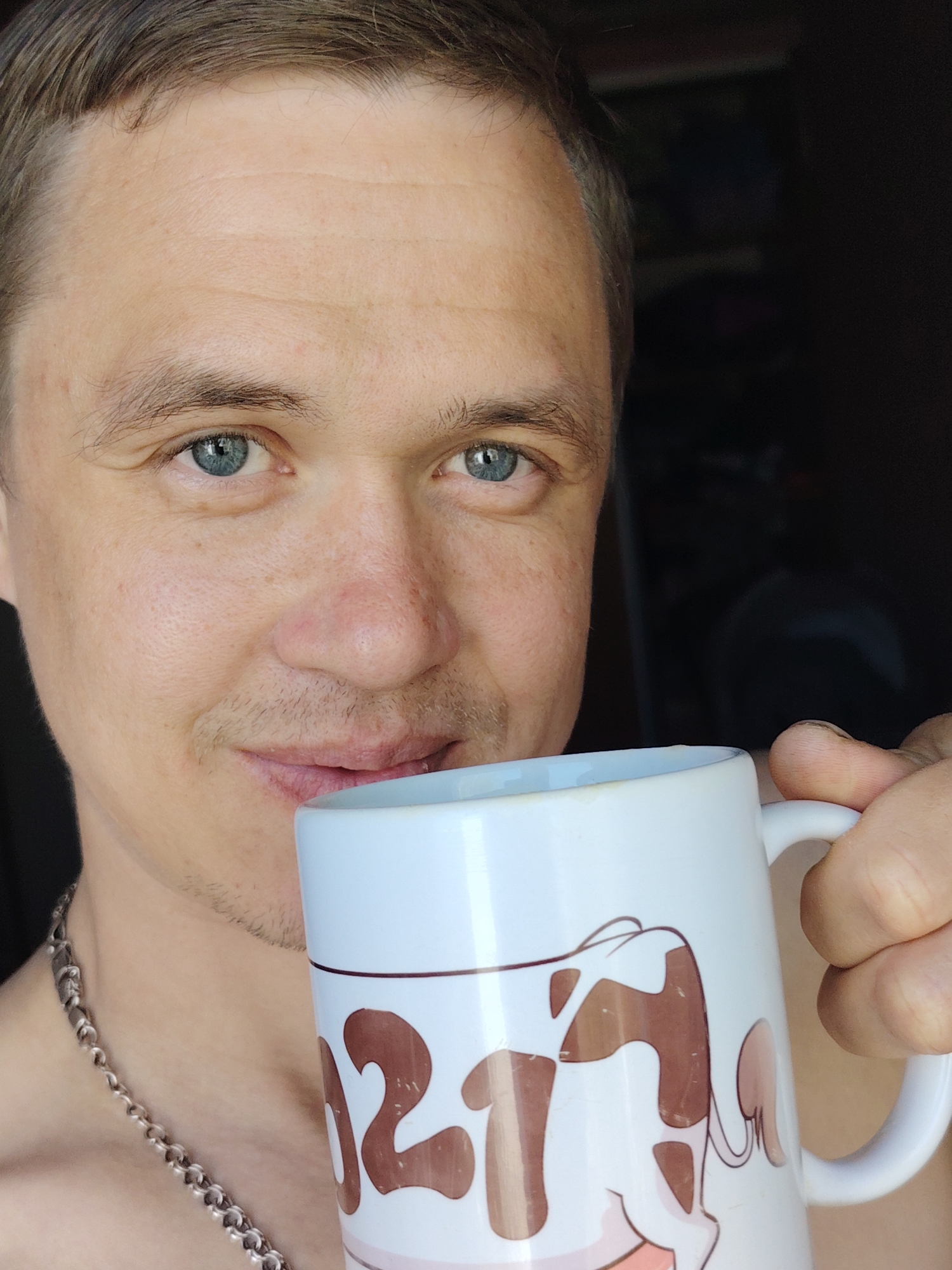 A close-up, medium-shot photograph shows a fair-skinned man with short brown hair and blue eyes holding a white mug with a cartoon cow design. He has a slight mustache and wears a silver chain necklace. The background is dark, suggesting an indoor setting. The exact location is unknown and cannot be determined from the image.