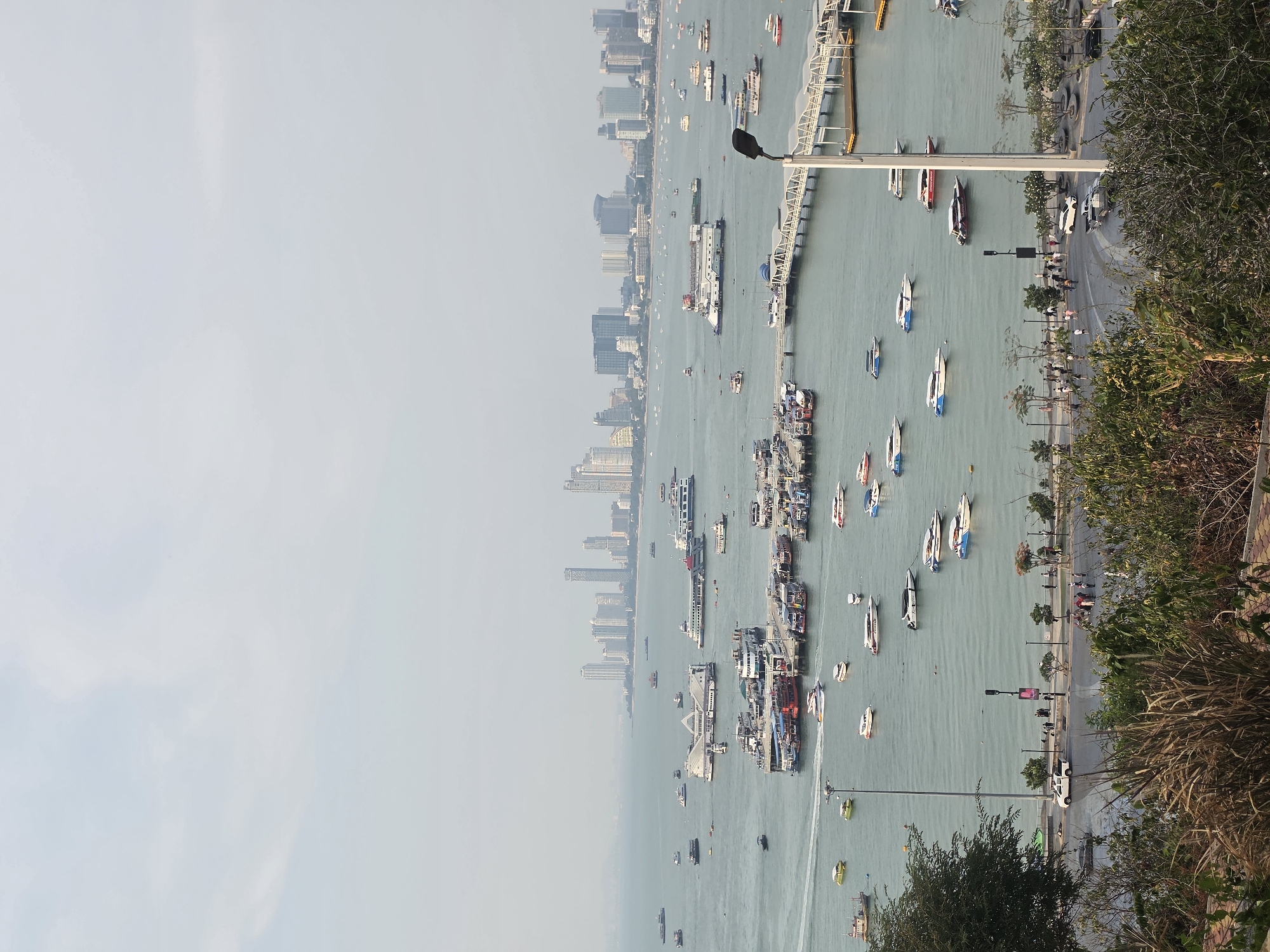 This is an aerial view of Pattaya City, Thailand, with a coastal city skyline stretching along the left side of the image. The main subject is a body of water, likely the Gulf of Thailand, filled with numerous boats and ships of varying sizes. A long pier or walkway extends into the water, supporting streetlights. Adjacent to the water is a road with pedestrians and parked vehicles. The right side of the image is dominated by vegetation, including trees and shrubs, from an elevated vantage point. The sky is overcast, with a hazy or smoggy appearance.