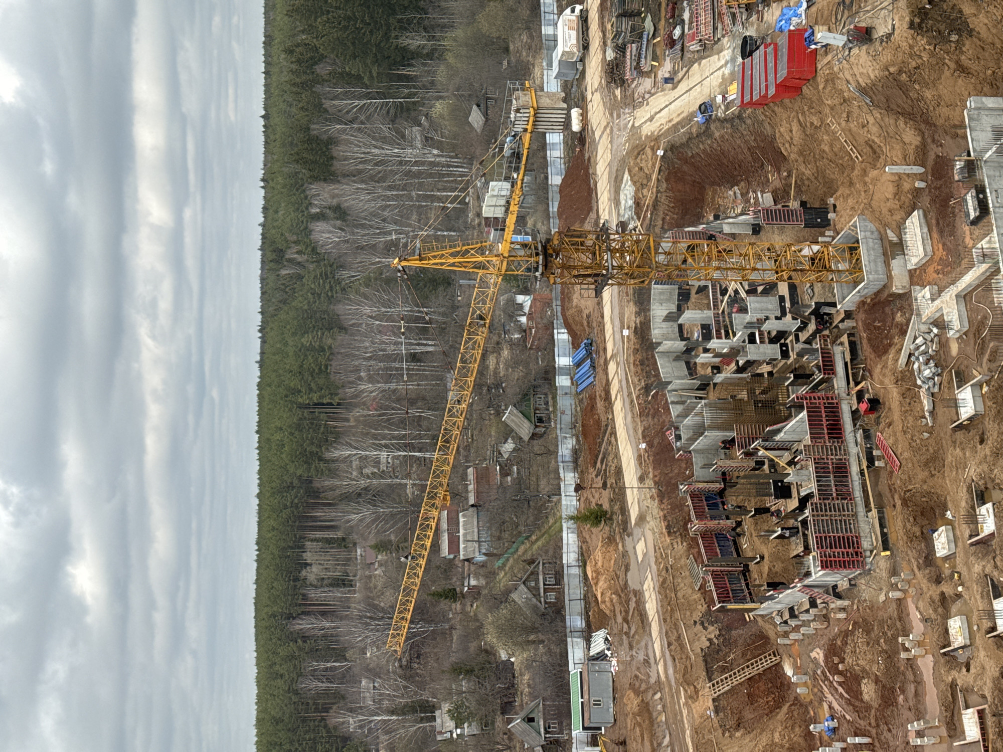 This aerial or high-angle image captures a large construction site in Izhevsk, Russia, situated at the edge of a dense forest under a uniformly overcast sky. The primary subject is a prominent yellow tower crane, its long arm extending over the active building area. The construction zone features numerous concrete foundations, partially erected structures with rebar, and extensive red and metal formwork for pouring concrete, indicating a multi-unit development. The ground is a mix of excavated earth, dirt roads, and areas covered with construction debris and materials such as stacked planks, concrete blocks, and blue tarps. Several deep pits and trenches are visible, suggesting significant groundwork.
Beyond the immediate construction activity, a sprawling forest dominates the left side of the frame, comprising both dark green coniferous trees and leafless deciduous trees, suggesting early spring or late autumn. Scattered within this natural landscape and along its edge are several small, older-looking houses with simple roofs, contrasting with the scale of the new development. A white truck is visible on a dirt path near the construction materials. The weather is cloudy and grey, consistent with daytime. No clearly legible text is discernible on any objects or structures within the image.