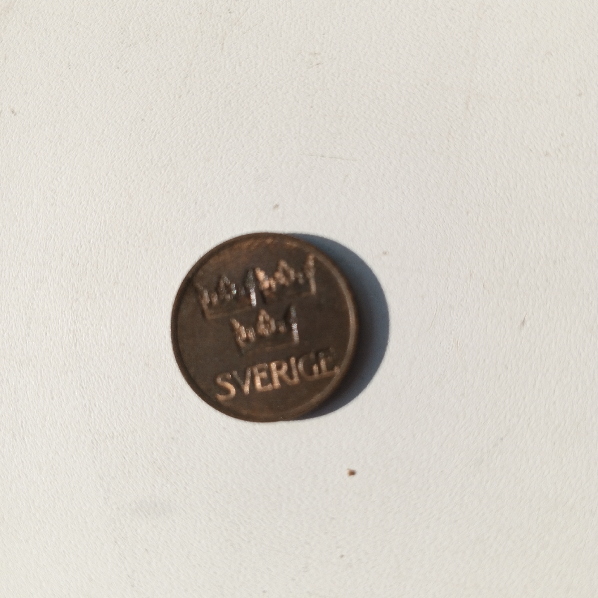 An overhead, medium shot captures a worn, copper-colored coin on a textured white surface. The coin is slightly off-center to the left and displays a prominent shadow to its right. Embossed on the coin's surface are two crowns above the word "SVERIGE". The white surface appears to be a textured wall or countertop. There are no people or any other discernible objects or elements in the frame. The scene is devoid of any specific location markers that would indicate Sayanogorsk, Russia.