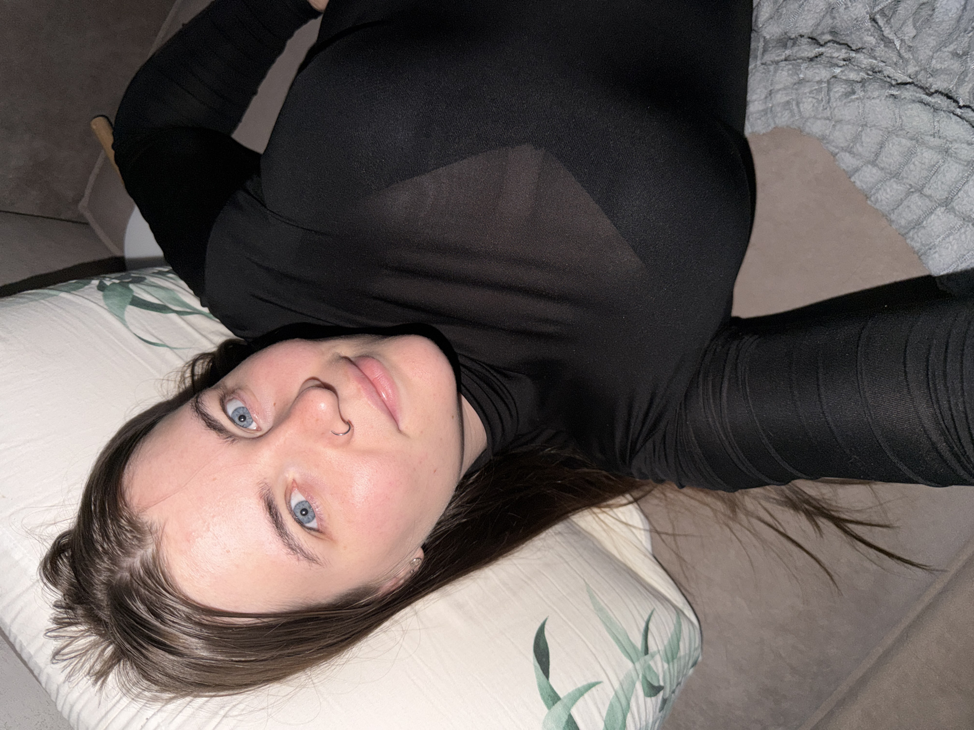 A young woman with blue eyes and long brown hair is lying on a couch, looking at the camera. She is wearing a black turtleneck shirt and grey pants. She has a nose ring and is wearing a small silver earring. The couch is covered with a patterned pillow and a grey textured blanket. The lighting is dim, suggesting it may be evening or indoors. The image is cropped to show the woman from the chest up. There is no indication of the location in Chaikovsky, Russia.