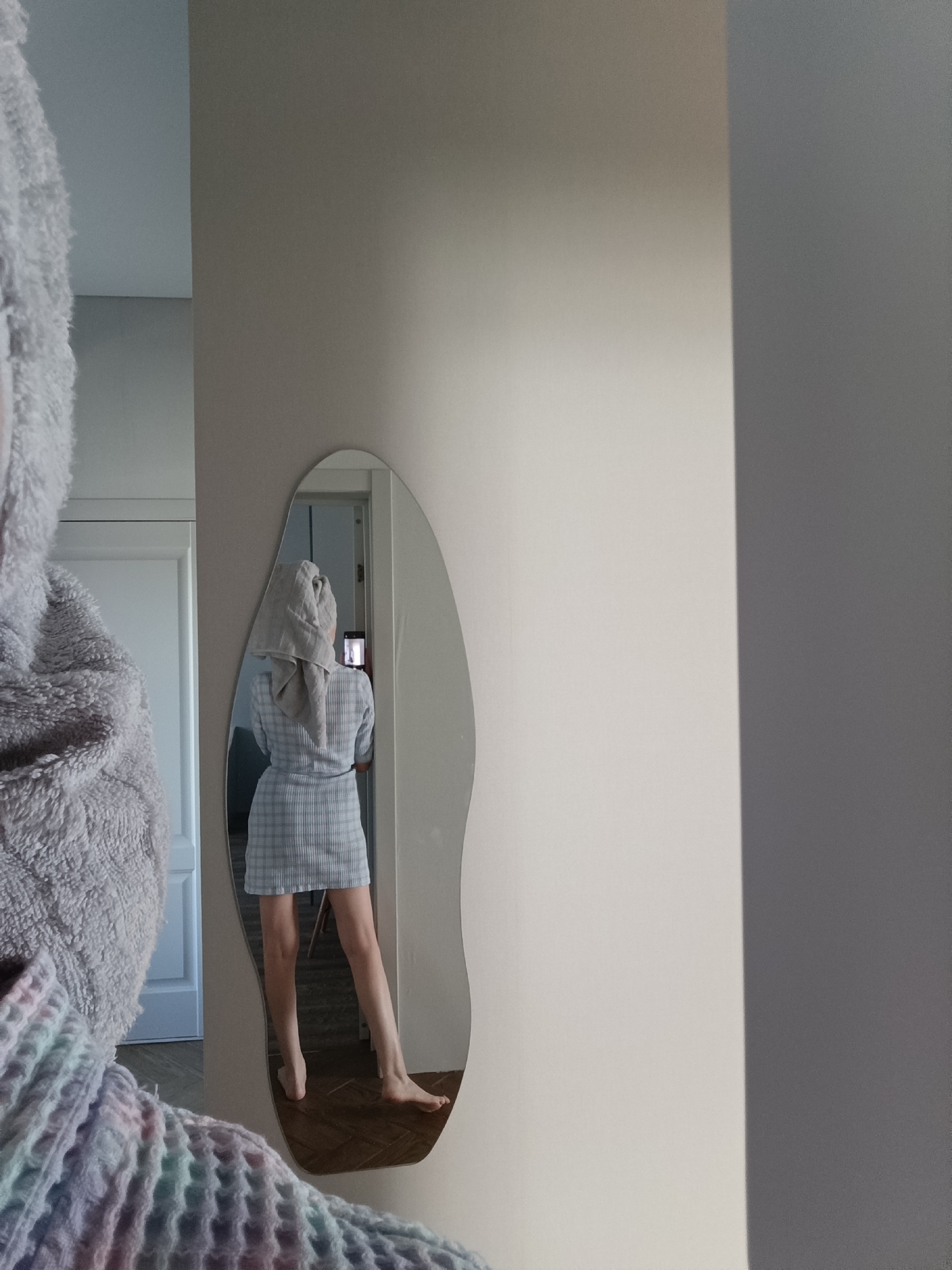 The image displays an indoor scene from a first-person perspective, capturing a mirror selfie. The photographer is partially visible on the left, out of focus, wearing a grey, plush-textured garment, with a multicolored, waffle-knit material draped over the left shoulder. A full-length, irregularly shaped mirror is mounted centrally against a light, neutral-toned wall.
Within the mirror's reflection, a female individual is observed from behind. She is dressed in a light blue and white plaid, short-sleeved mini-dress, and has a light grey towel wrapped around her head in a turban style. She holds a dark-colored smartphone in her right hand, extended to photograph her reflection. Her bare legs are visible from mid-thigh down, and her bare feet rest on a wooden floor featuring a herringbone pattern.
In the background of the reflection, to the left of the reflected subject, a white door frame and a closed white door are visible. Natural light illuminates the scene, casting a prominent vertical shadow on the right side of the image. No discernible visual information identifies a specific location within the city of Chaikovsky, Russia.