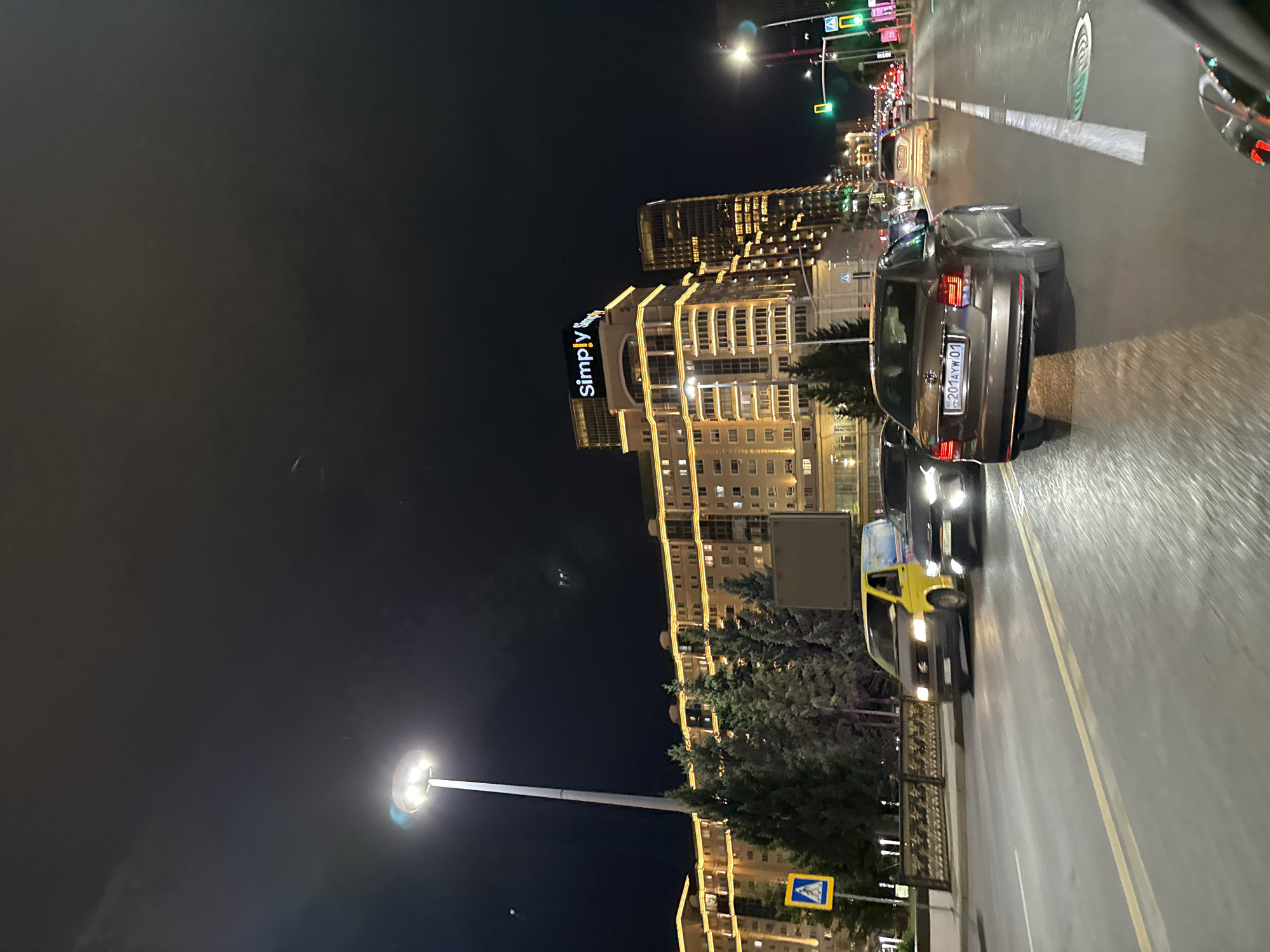 A nighttime street scene in Astana, Kazakhstan, features a multi-lane road with moderate traffic. The foreground shows the rear of a dark-colored passenger car with a license plate reading "201AYW 01". To the left, a smaller, yellow van is visible, followed by another dark-colored vehicle. In the background, a large, illuminated building complex with a "Simply" sign at its top dominates the skyline. The building is adorned with decorative yellow lighting. Adjacent to it is a taller, modern skyscraper with illuminated windows. Traffic lights are visible, with one showing green. Trees line the median of the road, and a pedestrian crossing sign is present on the left side. The street appears wet, possibly from recent rain, reflecting the streetlights and vehicle lights. The sky is dark and cloudy.