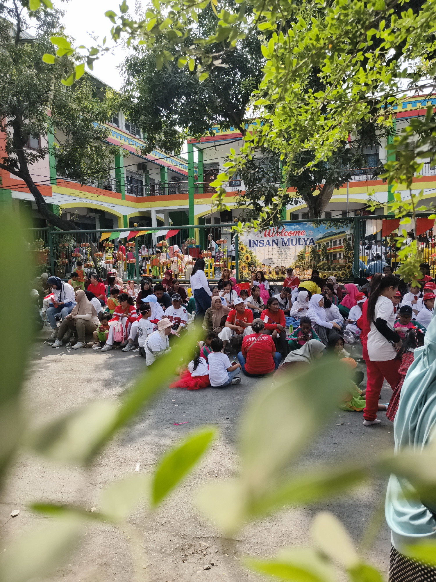 In the city of Petiken, Indonesia, a gathering of people is observed in front of a brightly colored building, likely a school, with a banner that reads "Welcome INSAN MULYA". The building features multiple stories, balconies, and vibrant stripes of yellow, orange, red, and green along its facade. A green fence separates the crowd from the building. A large group of individuals, a mix of adults and children, are seated on the ground, facing towards the building. Many are wearing red and white clothing, suggesting a celebration or event. Some children are dressed in festive outfits, including red tutus. In the foreground, out-of-focus green leaves and branches partially obscure the view of the scene. The ground appears to be a paved area, possibly a courtyard or playground. The overall atmosphere suggests a community event or school function.