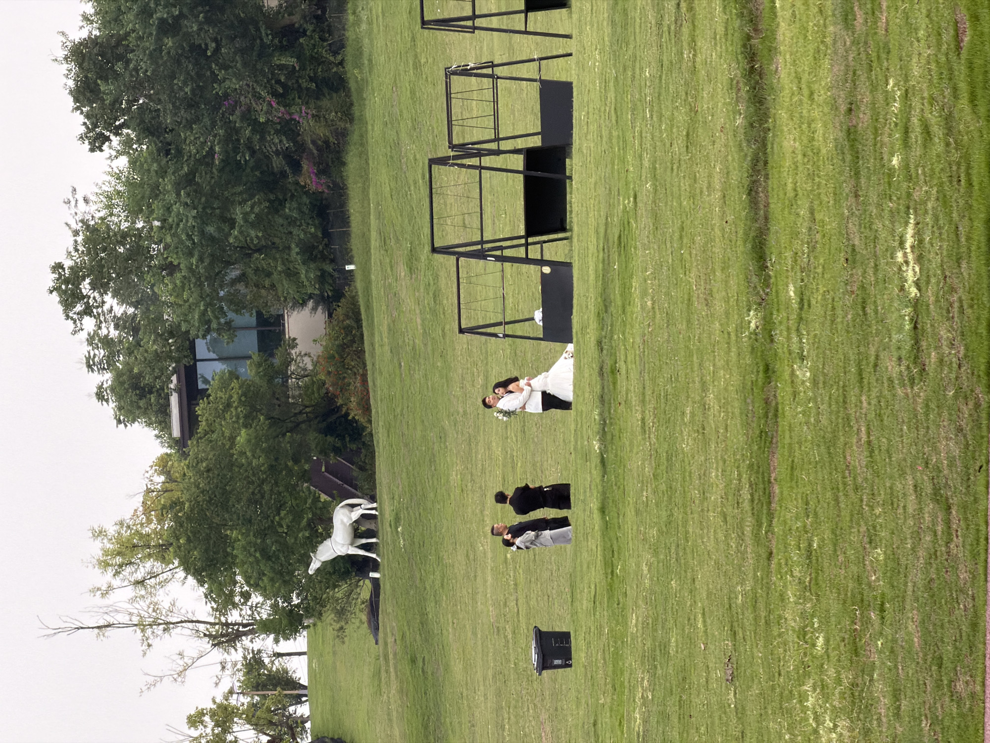 In a vast, open grassy field in Jiangjin District, Chongqing, China, a wedding photoshoot is taking place. A bride in a white gown and a groom in a white suit are posed embracing each other, with the groom holding a bouquet of white flowers. To their left, three individuals, two men in black attire and one woman in a grey outfit, stand together, likely part of the wedding party or photographers. Further to the left, a black trash can sits on the grass. In the background, a white statue of a horse with its head raised stands on a small incline. Behind the horse, a modern building with large windows is partially visible, surrounded by lush green trees and some blooming pink flowers. Several black metal structures, possibly swings or decorative elements, are also scattered across the field, adding to the scenic, outdoor setting. The sky is overcast, suggesting a muted natural light for the event.