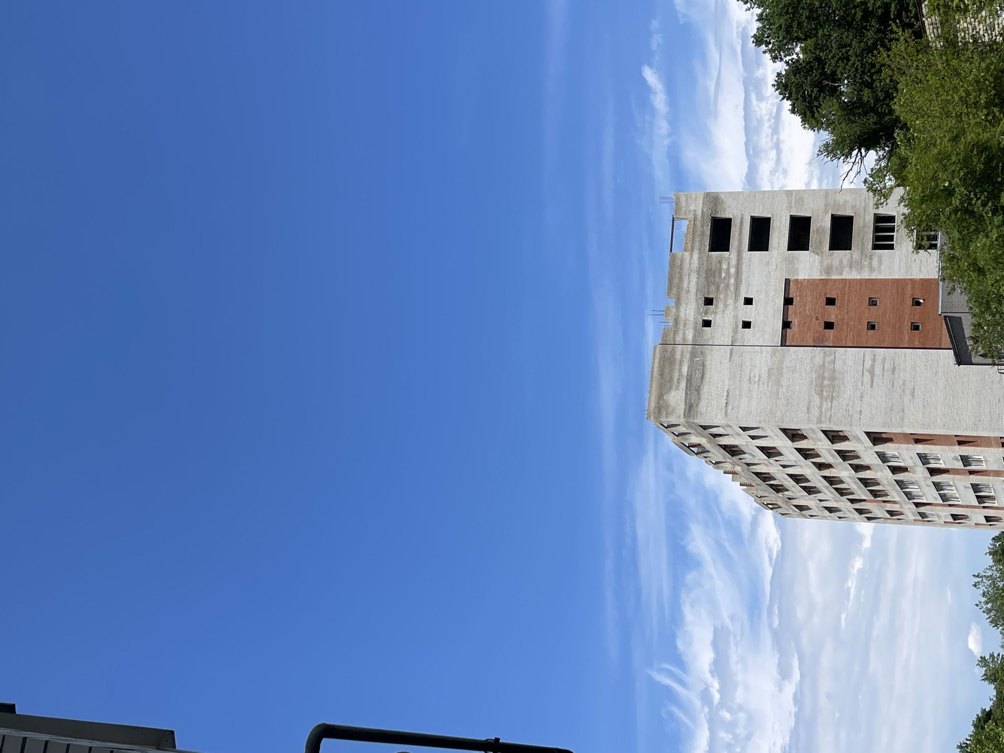 This media file displays a partially constructed multi-story building under a bright blue sky with scattered white clouds. The building appears to be made of concrete and brick. The concrete sections have numerous rectangular openings, suggesting unfinished window frames. A portion of the building is clad in reddish-brown brick, also with square openings. Some rebar is visible extending from the top of the structure, indicating ongoing construction. Green trees are visible at the top right of the frame, partially obscuring the building. The orientation of the image is rotated 90 degrees counter-clockwise. The clear sky and sunlight suggest it is daytime, likely a pleasant weather day. There is no visible text in the image. The scene appears to be an urban or suburban setting where construction is underway.