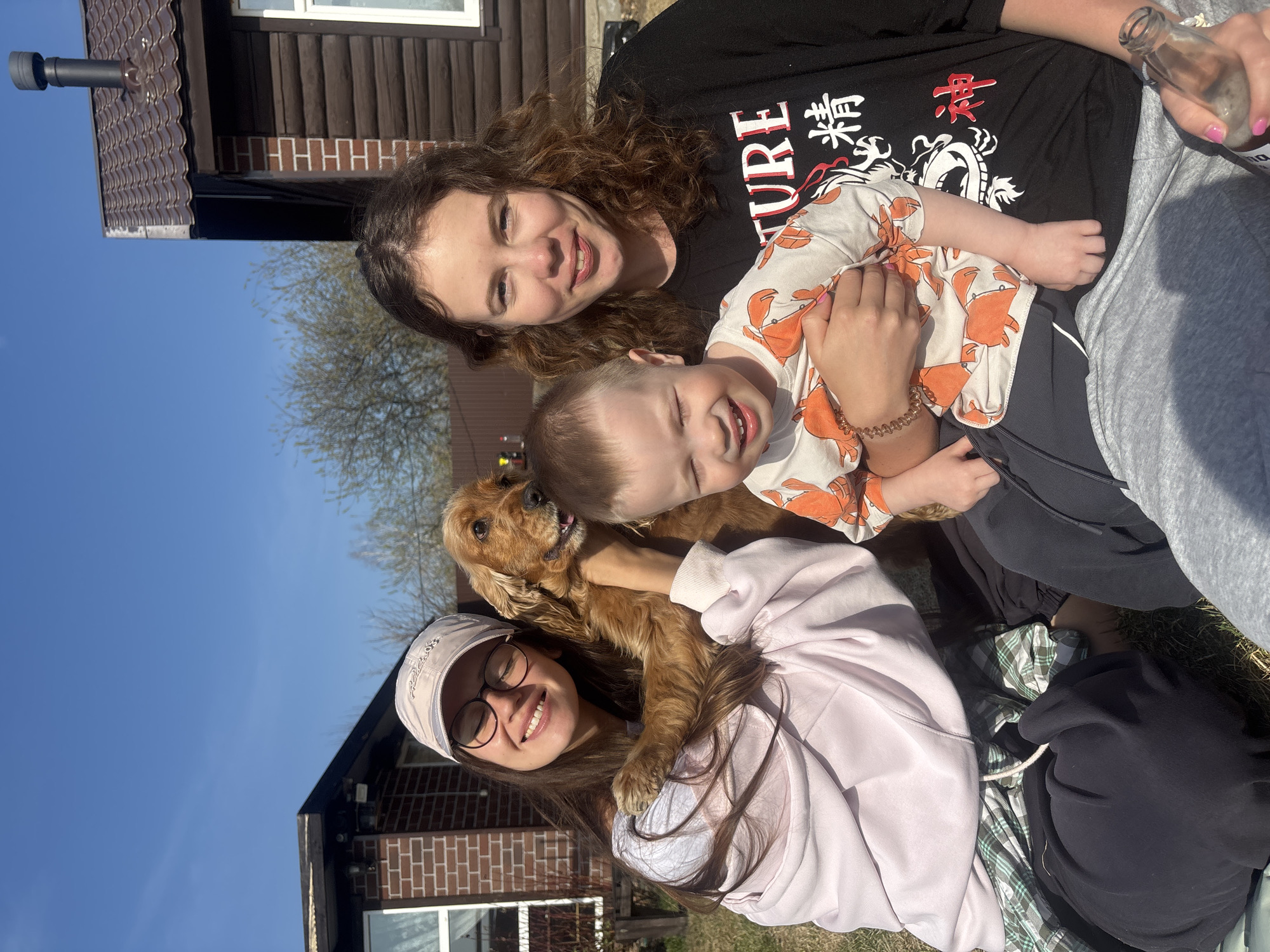 The image shows three people and a dog in an outdoor setting. A young child wearing a light-colored shirt with orange crab patterns is being held by an individual in a black shirt with white and red writing and a dragon design. Next to them is another individual, possibly a woman, with curly brown hair. In the foreground, a person wearing a light pink hoodie and a baseball cap is holding a golden-colored dog. They all appear to be smiling or laughing. The background shows a wooden building with a brown roof and brick accents. The location appears to be outdoors, possibly a yard or garden. The sky is blue. No information is available confirming the location is in the city of Emel'yanovo, Russia.