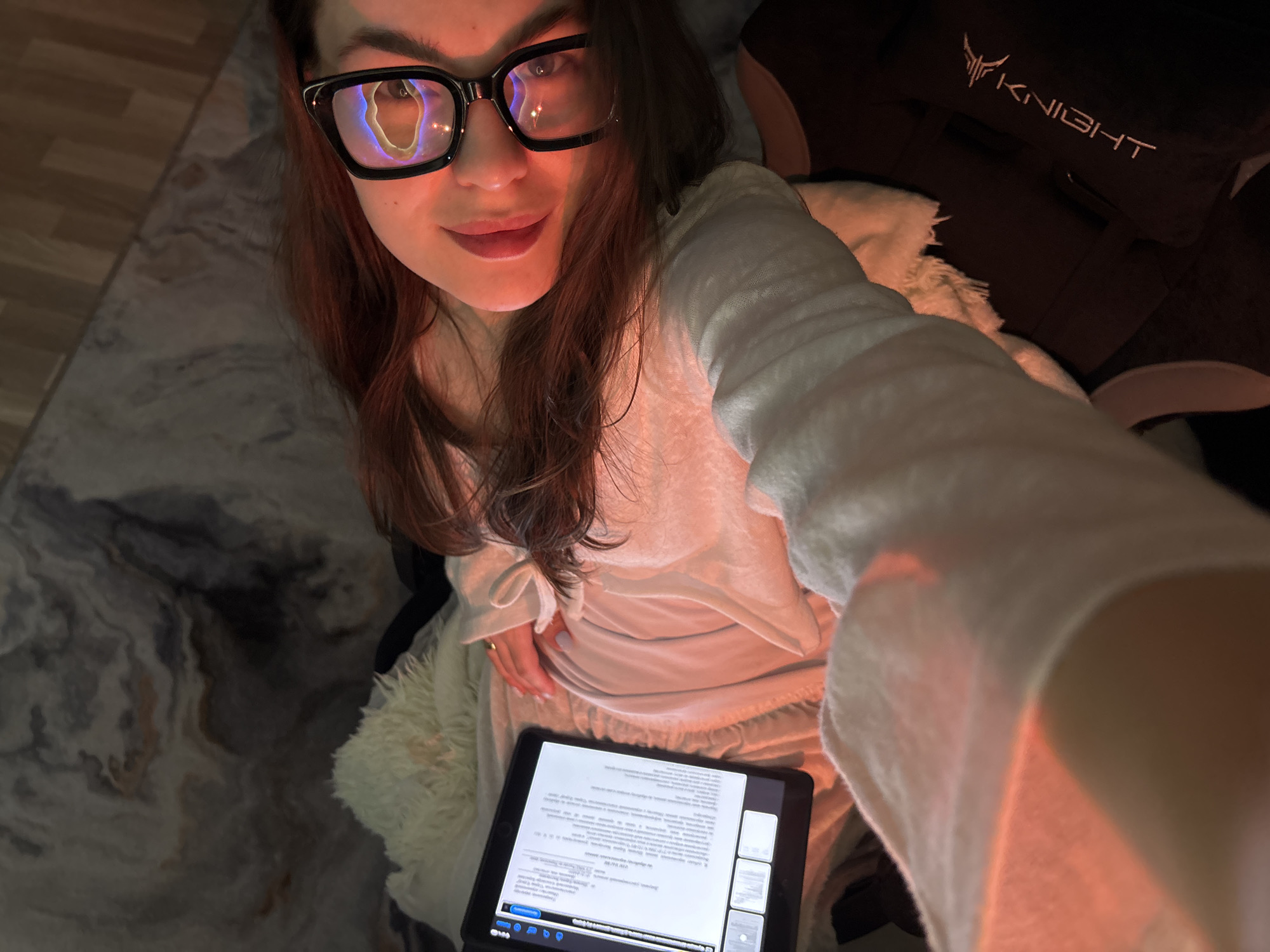A woman with long brown hair and wearing large black-framed glasses is taking a selfie. The reflection in her glasses suggests she is indoors, illuminated by a screen. She is wearing a light-colored, possibly white, long-sleeved pajama top. She is holding a tablet in her hands displaying a document with text. She appears to be sitting or reclining, with part of a black gaming chair with "KNIGHT" embroidered on it visible in the background. There is a fluffy white blanket or throw around her. The floor has a marbled pattern. The city of Chaikovsky, Russia, is not discernable from the image.