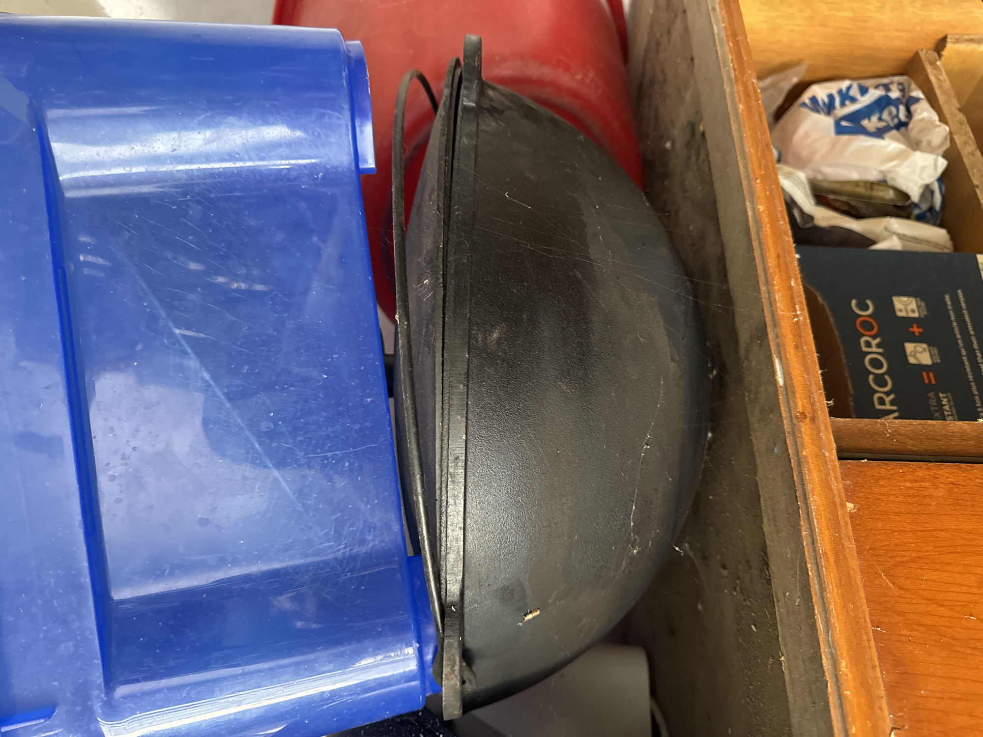 In a dimly lit interior space, possibly a storage area or workshop, a black cast iron pot with a lid sits on a wooden shelf. The pot is positioned in front of a large, bright blue plastic bin and partially obscured by a red container behind it. The environment appears dusty, with visible cobwebs on the shelf and the pot. In the lower right portion of the image, a wooden cabinet is visible, with a partially opened white plastic bag containing items and a blue and white box labeled "ARCROC" placed on a shelf below. The overall scene suggests a cluttered and utilitarian setting. There is no indication of the city of Rozdilna, Ukraine, within the image content.